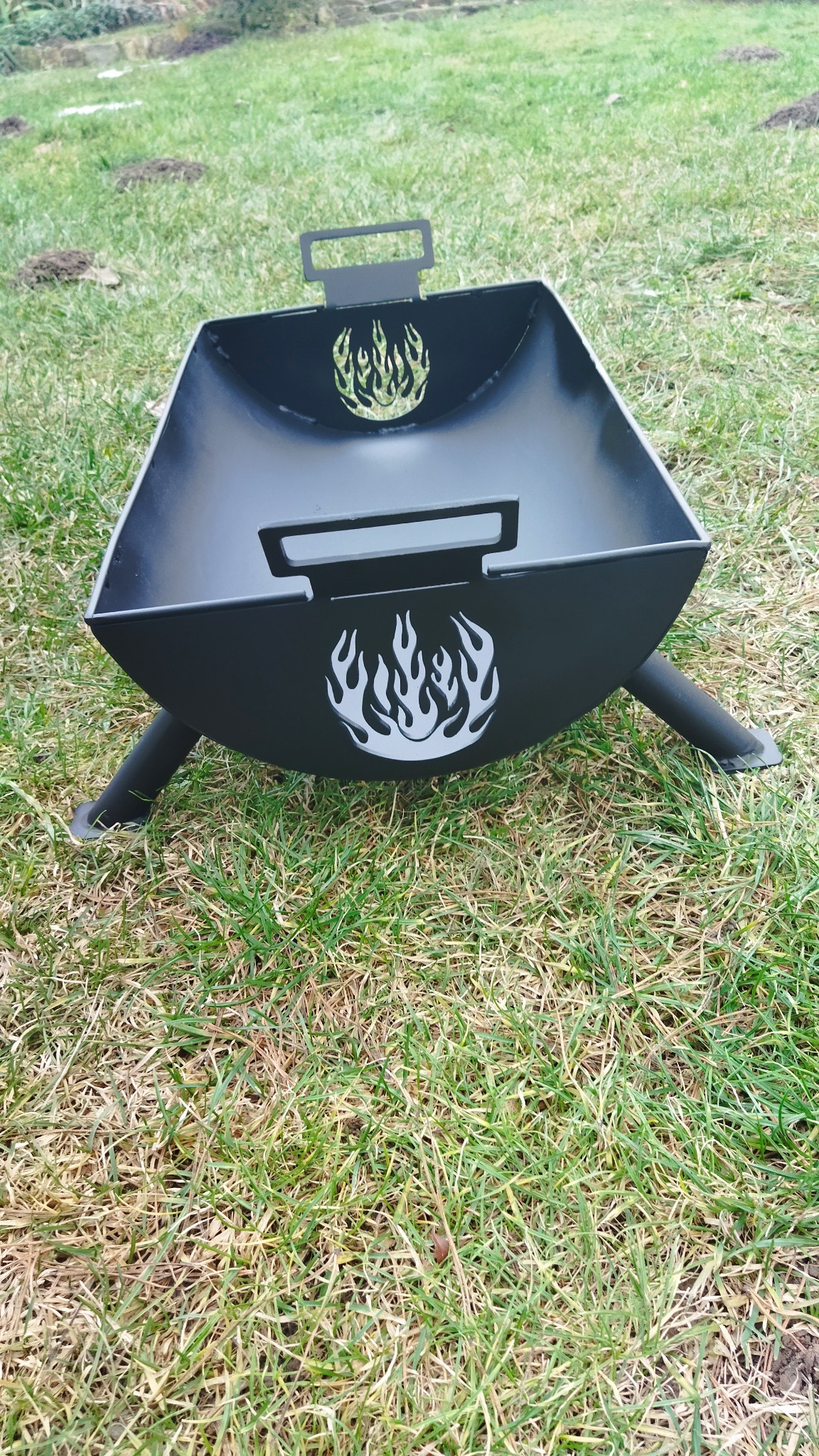 Czarny, metalowy kosz na ognisko z motywem płomieni, stojący na trawie.