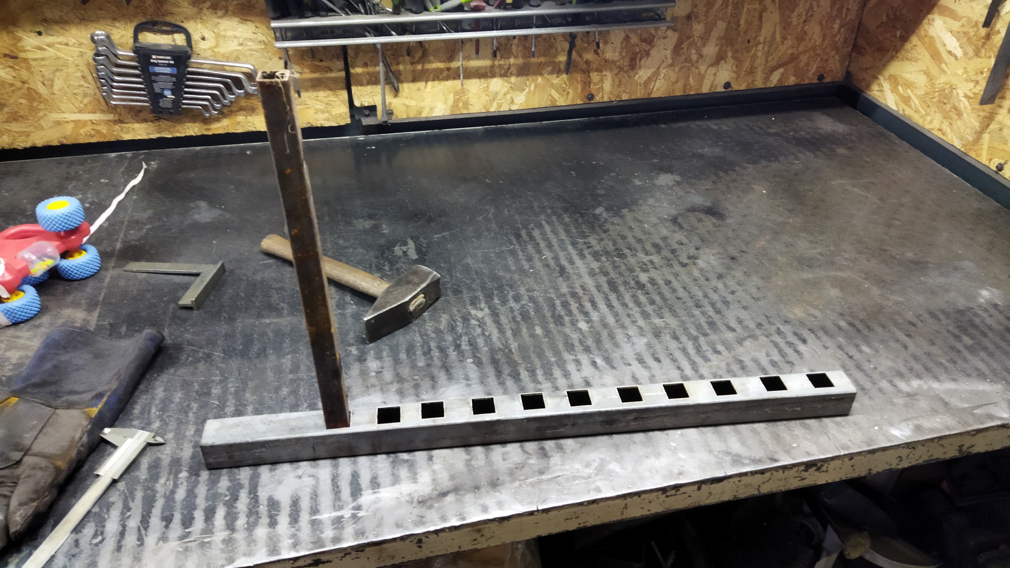 Metalowa konstrukcja z rur kwadratowych, z wyciętymi otworami, umieszczona na warsztatowym stole obok młotka, kątownika, suwmiarki i rękawic roboczych. W tle widoczne narzędzia na ścianie i zabawka.