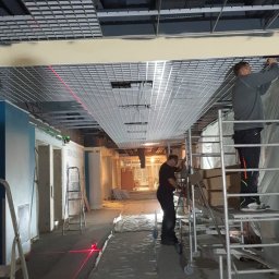 U.T. Construction - Montaż sufitu podwieszanego rastrowego w biurze, dwóch pracowników na rusztowaniach, widoczny laser krzyżowy wyznaczający poziom, zabezpieczona folią podłoga.