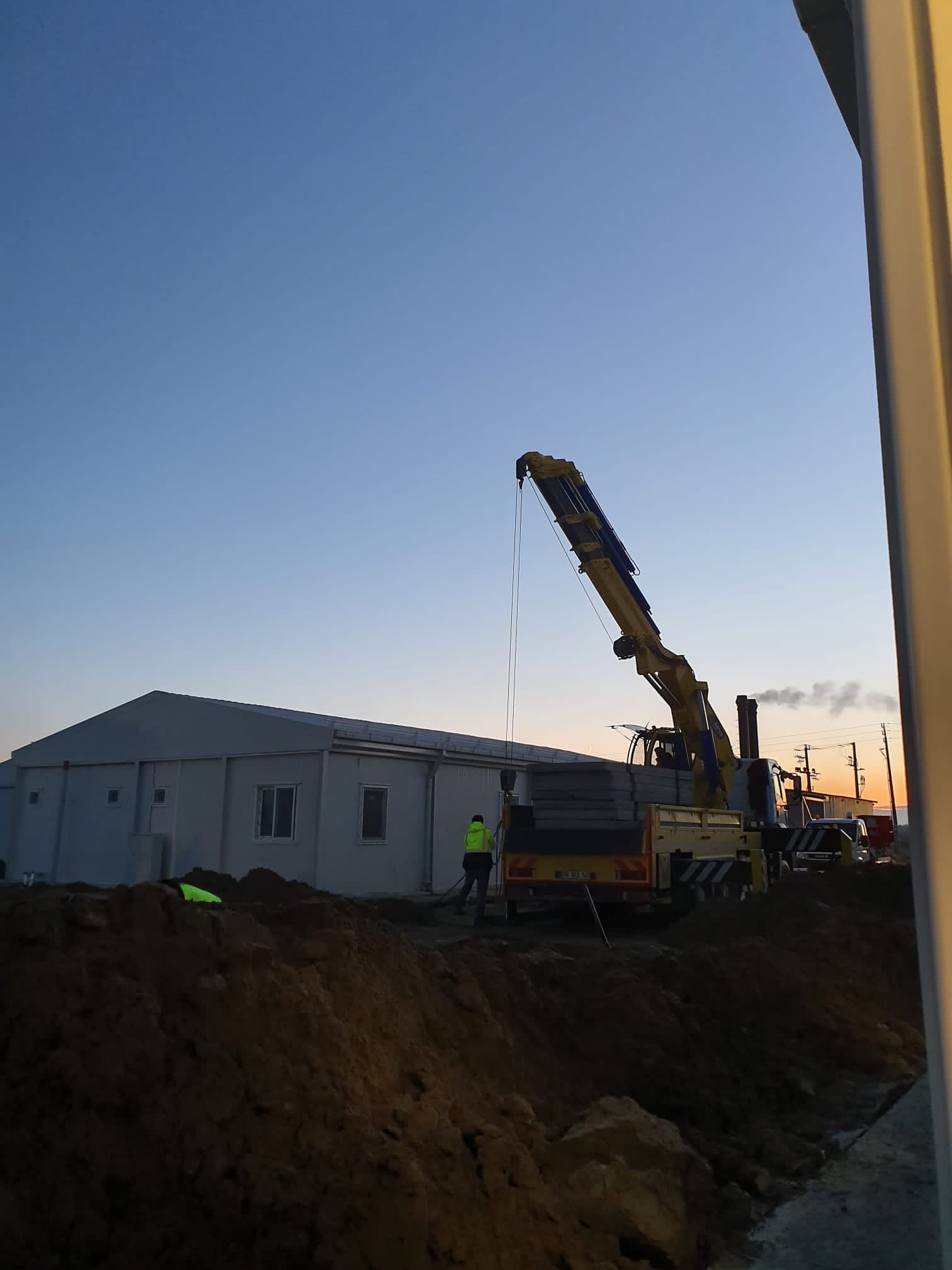 Żółto-niebieski dźwig samochodowy podnosi prefabrykowane betonowe płyty na tle szarego budynku modułowego i kopca ziemi o zmierzchu.