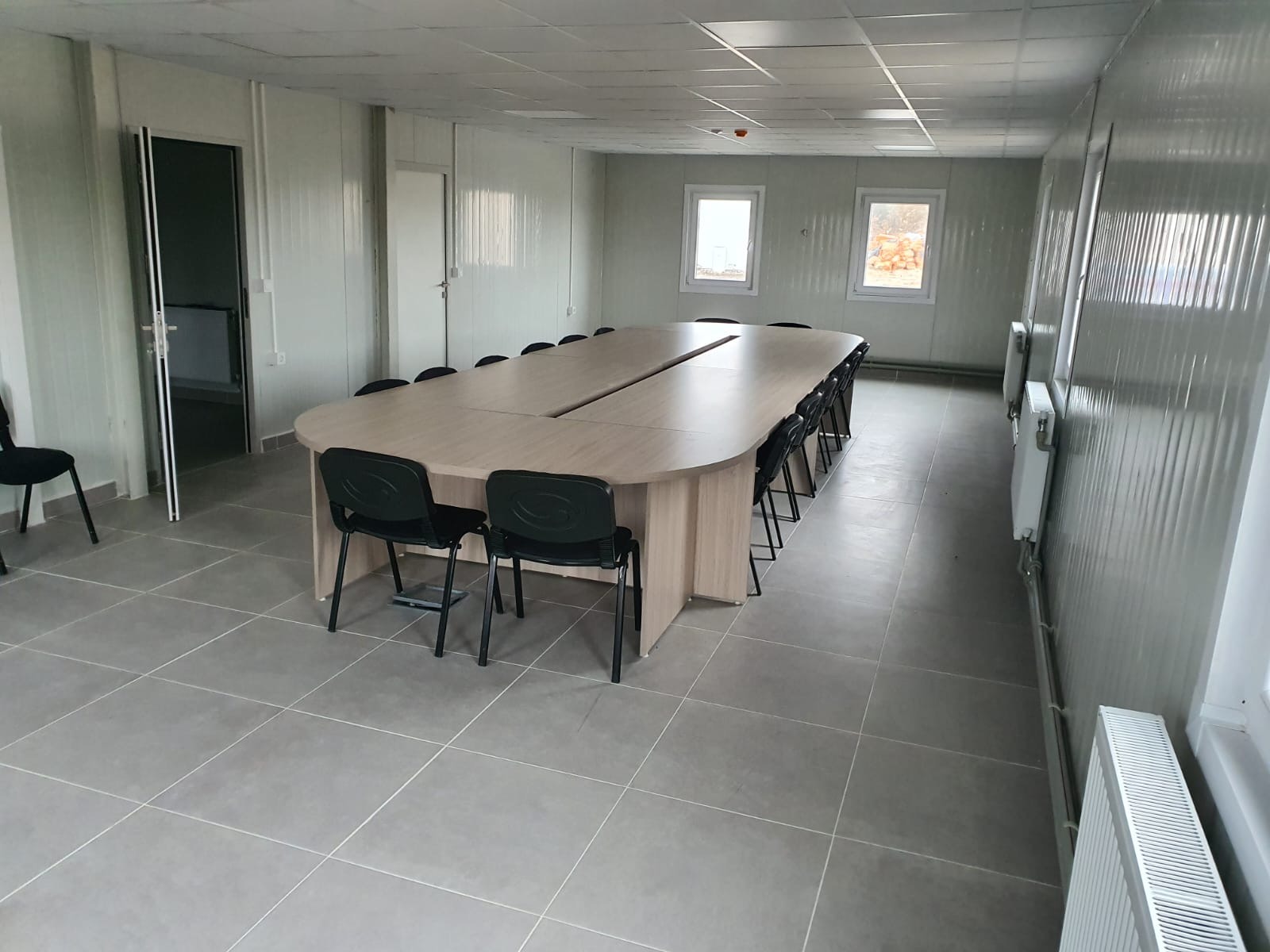 Wnętrze biura z długim stołem konferencyjnym i krzesłami, szare płytki podłogowe, białe ściany z blachy falistej i grzejniki pod oknami.