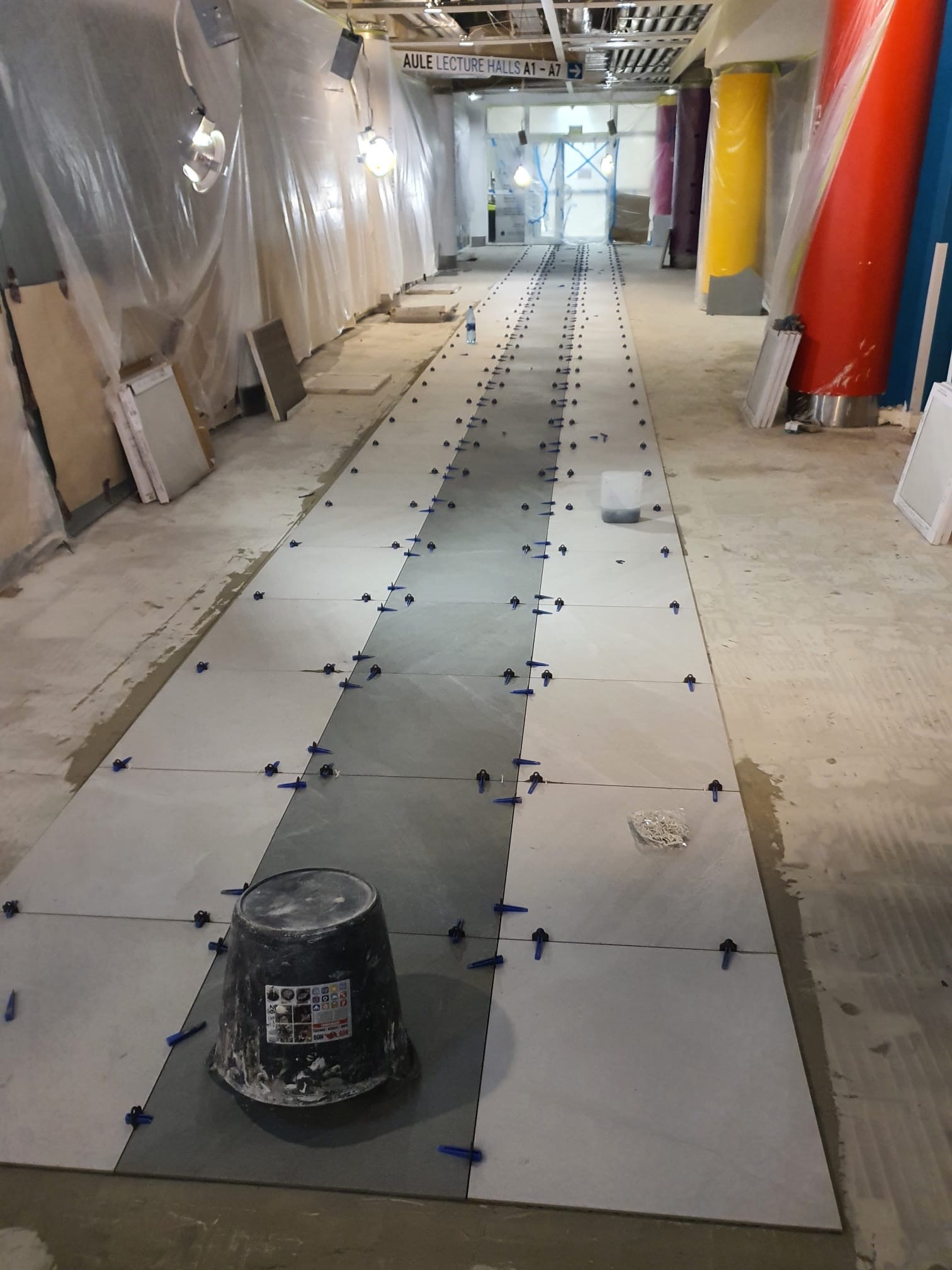 Układanie szarych płytek podłogowych w długim korytarzu z kolorowymi kolumnami, widoczne niebieskie klipsy poziomujące i wiadro z zaprawą.