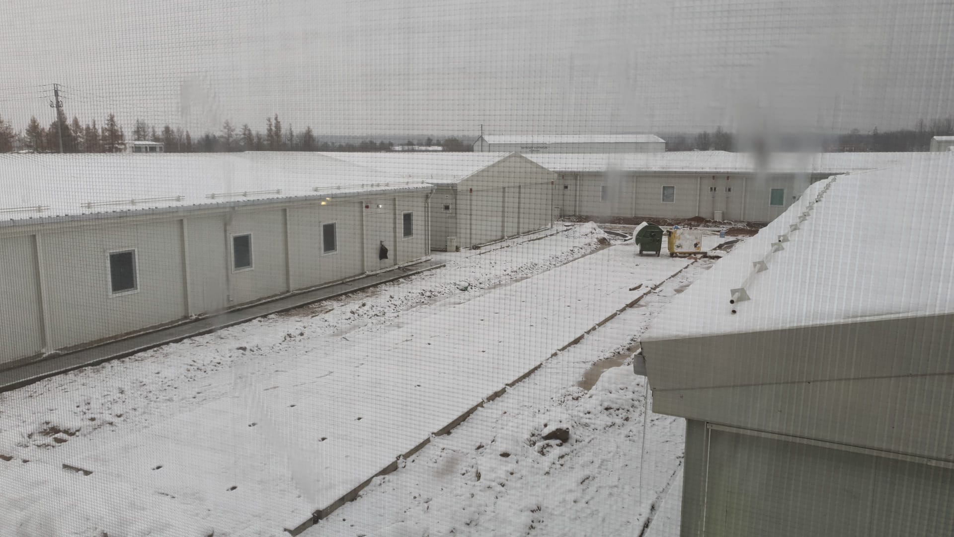 Widok z okna na modułowe budynki pokryte śniegiem, z widocznymi kontenerami na śmieci i fragmentem dachu budynku na pierwszym planie.