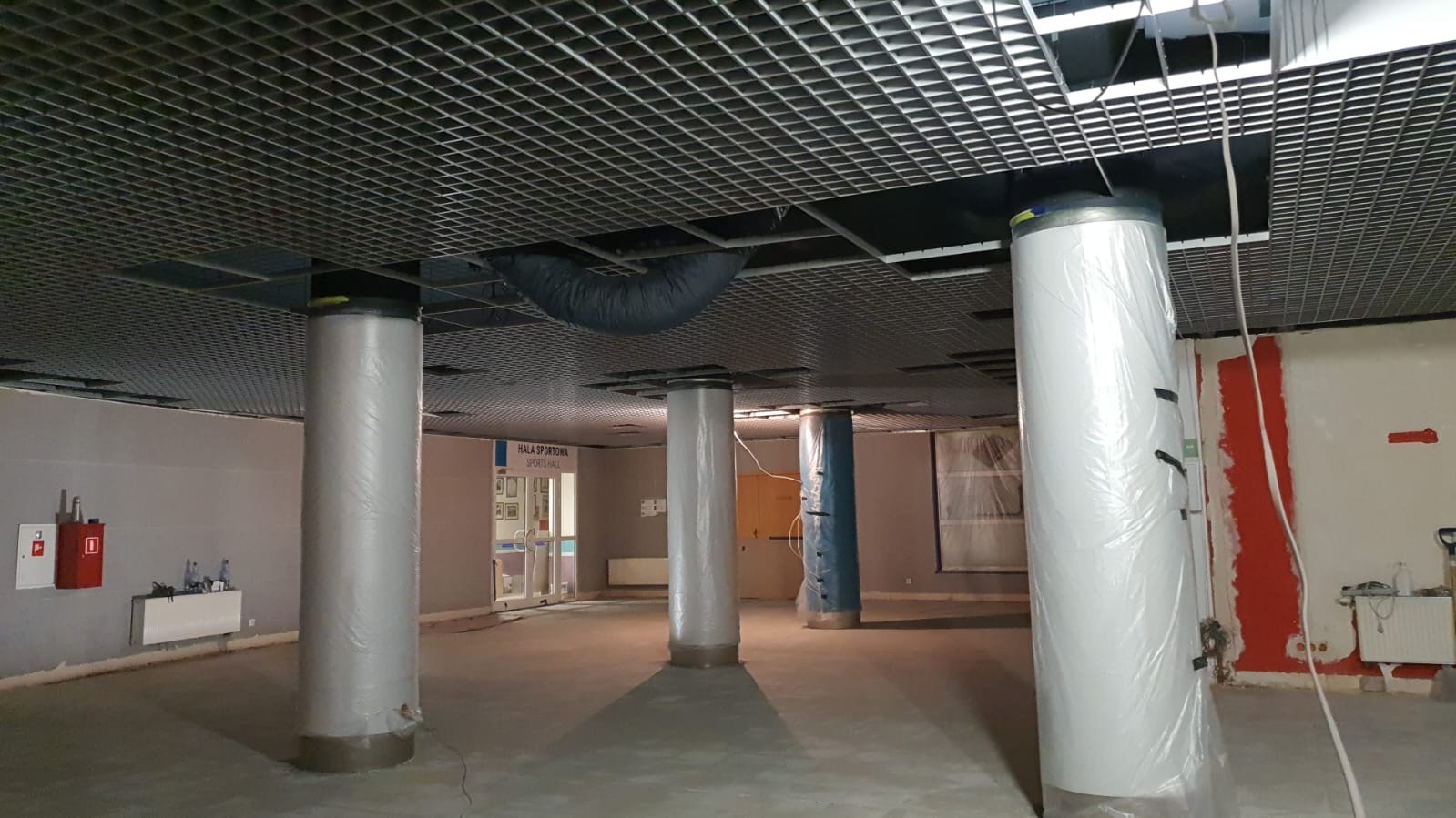 Wnętrze hali sportowej w trakcie prac remontowych: kolumny zabezpieczone folią, sufit podwieszany z kratką, widoczne elementy instalacji wentylacyjnej, ściany częściowo pomalowane na czerwono.