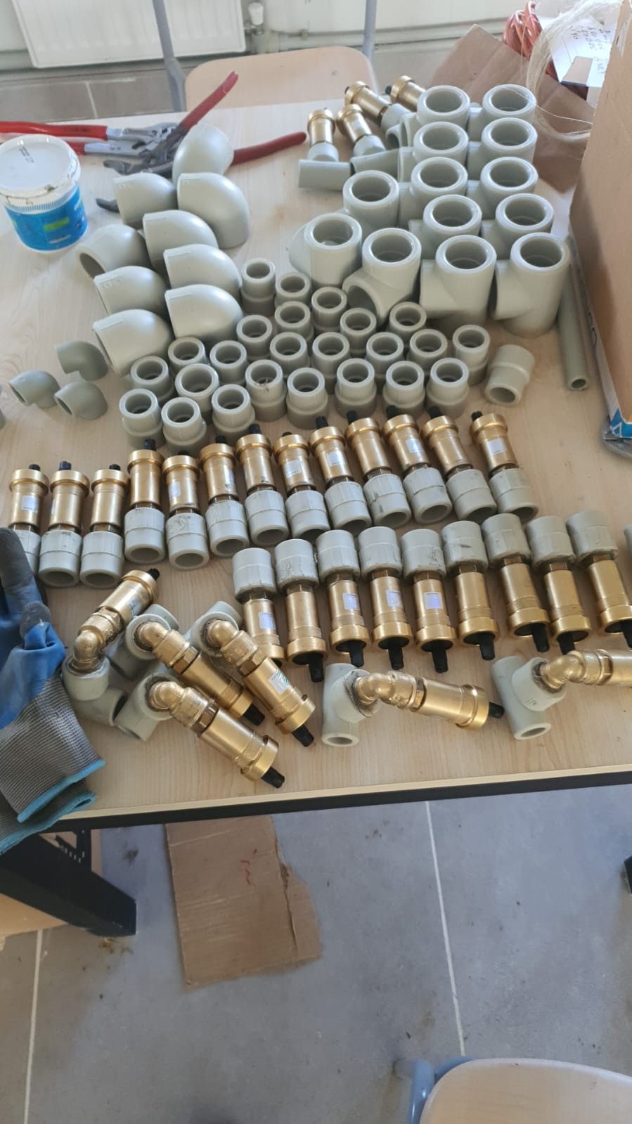 Różne złączki i kształtki instalacyjne z tworzywa i mosiądzu ułożone na stole, widoczne również narzędzia i rękawice robocze.