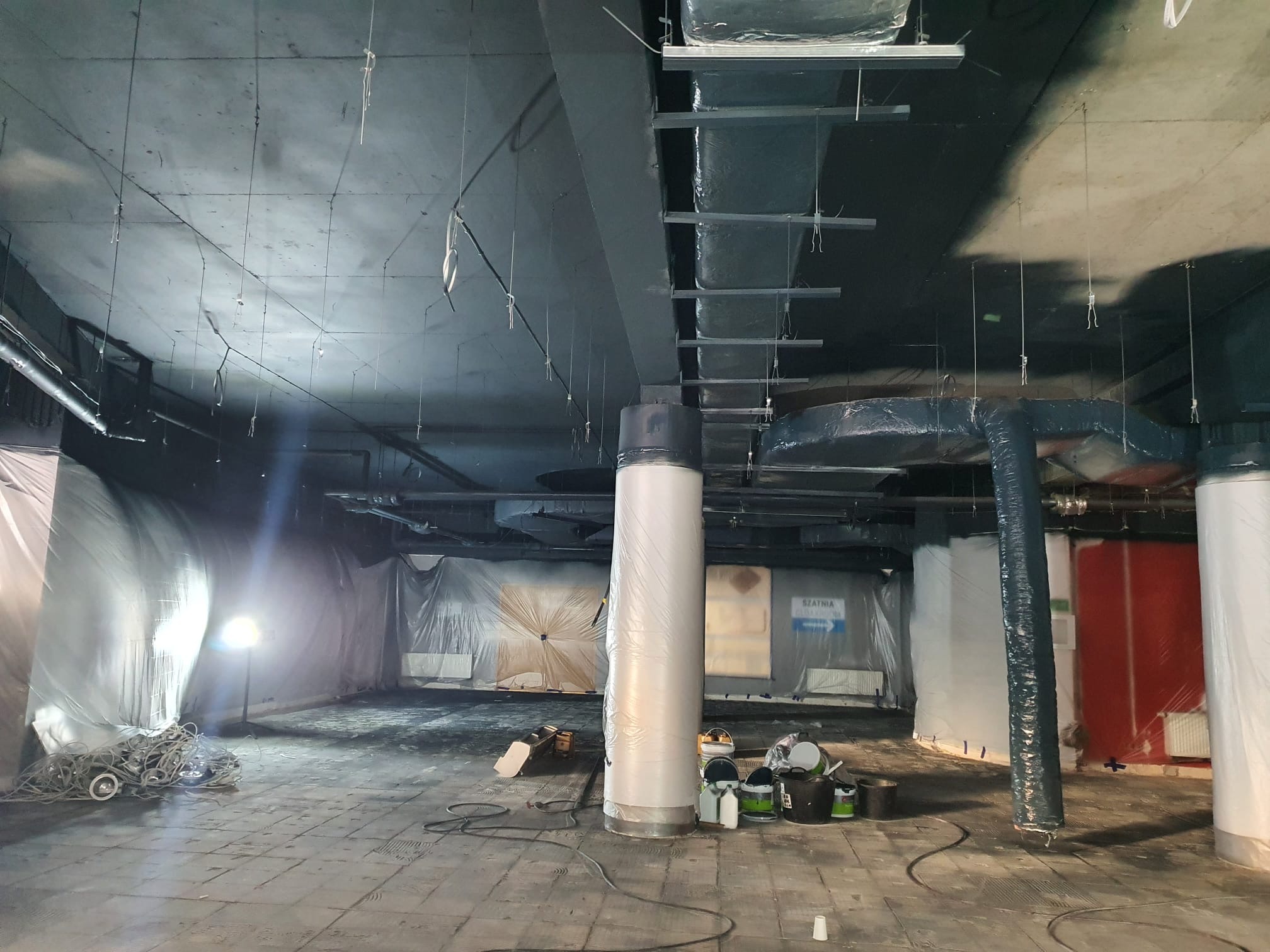 Wnętrze w trakcie przygotowania do prac wykończeniowych, z zabezpieczonymi folią ścianami i kolumnami, widoczne puszki z farbą i lampa halogenowa.