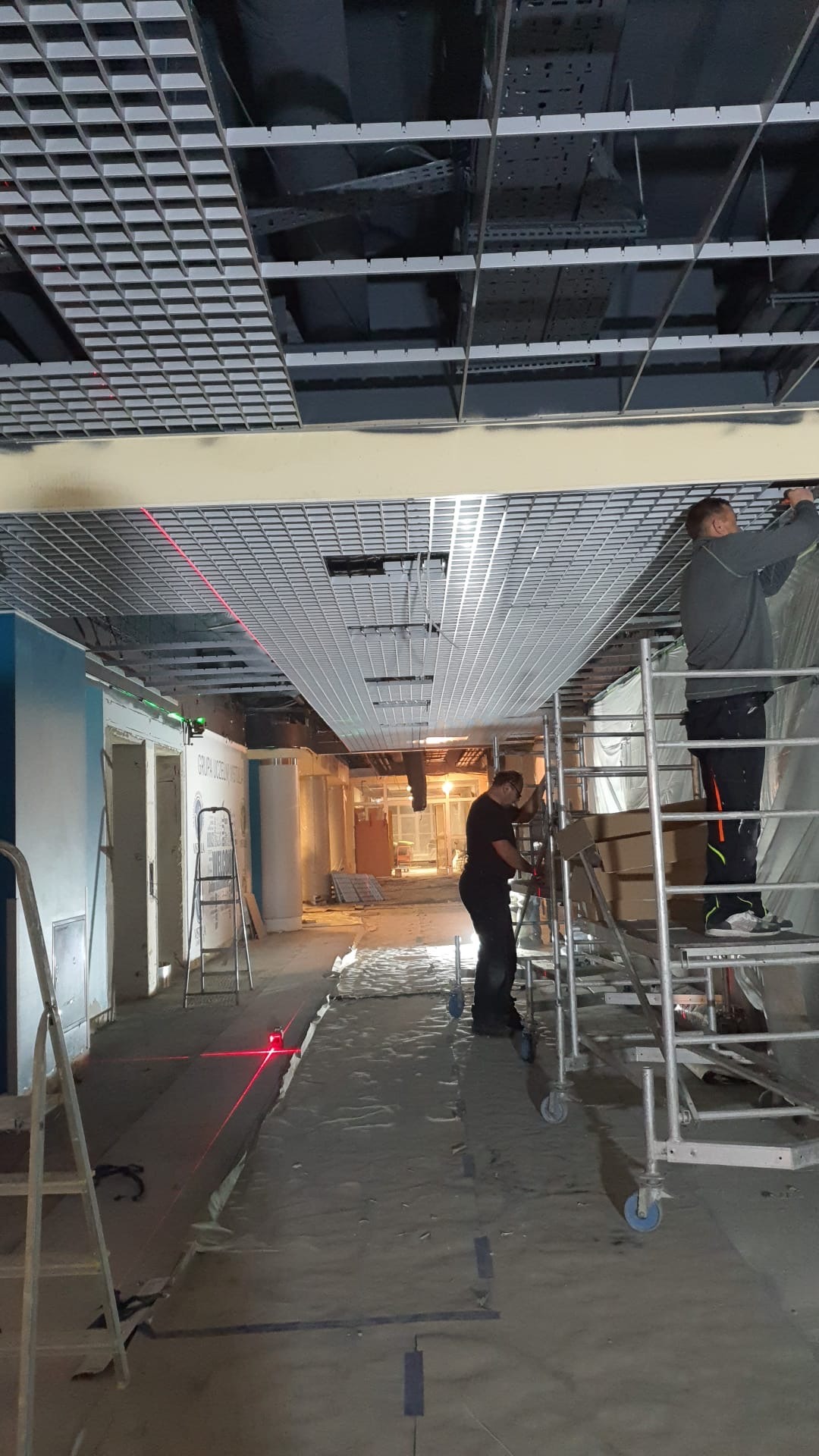 Montaż sufitu podwieszanego rastrowego w biurze, dwóch pracowników na rusztowaniach, widoczny laser krzyżowy wyznaczający poziom, zabezpieczona folią podłoga.