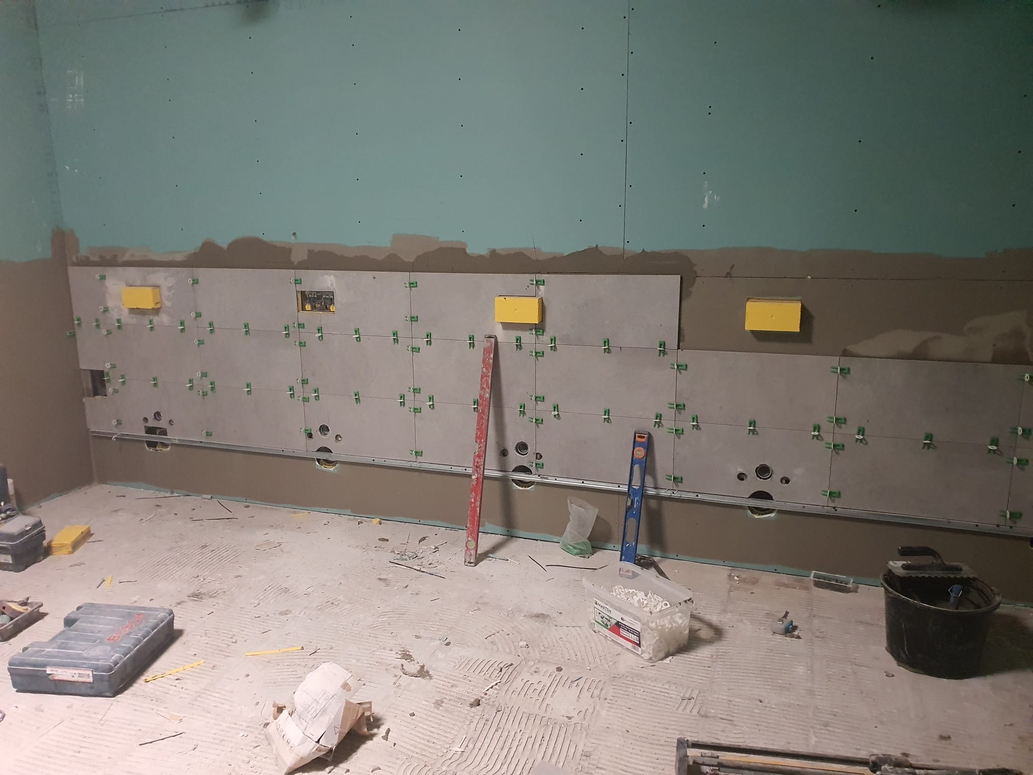 Ściana w trakcie układania płytek ceramicznych, widoczne poziomice, krzyżyki dystansowe, puszki elektryczne i narzędzia na podłodze.