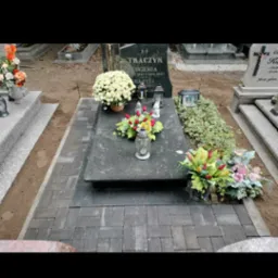 Grób z ciemnoszarą kostką brukową ułożoną wokół nagrobka, na którym stoją kwiaty i znicze.