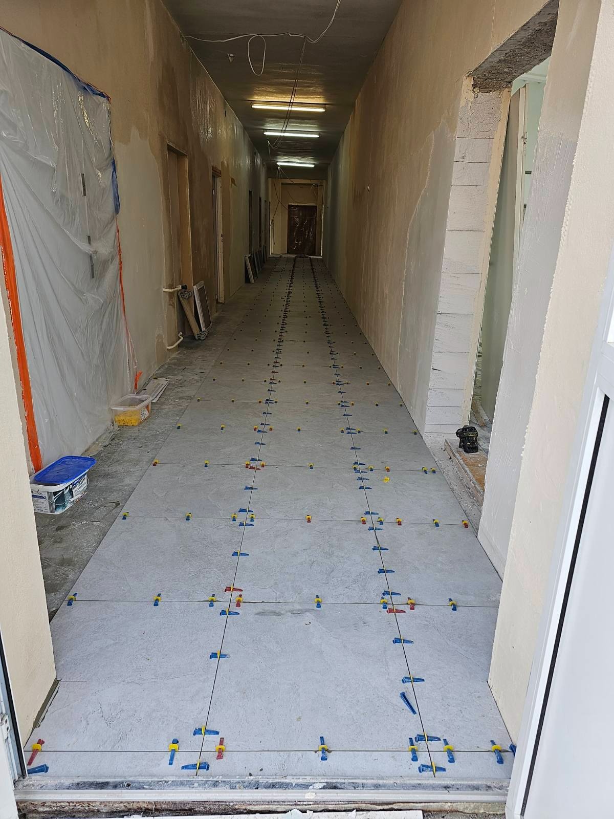 Długi korytarz w trakcie układania szarych płytek podłogowych, z użyciem klipsów poziomujących; ściany przygotowane do malowania, zabezpieczone folią ochronną.
