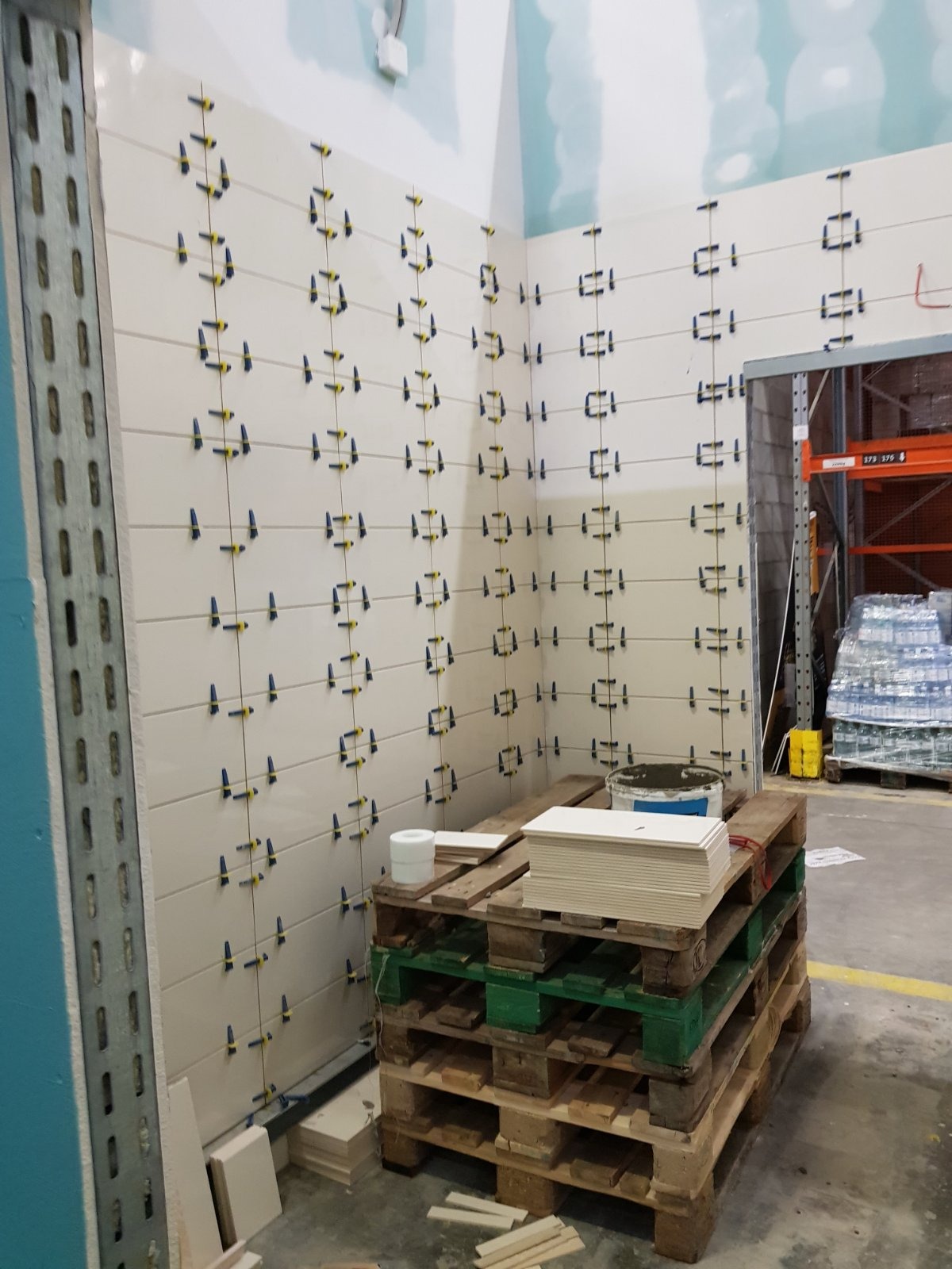 Świeżo ułożone, białe płytki ścienne z niebieskimi i żółtymi klipsami poziomującymi, ułożone na palecie z zaprawą i rolką taśmy malarskiej w tle magazynu.