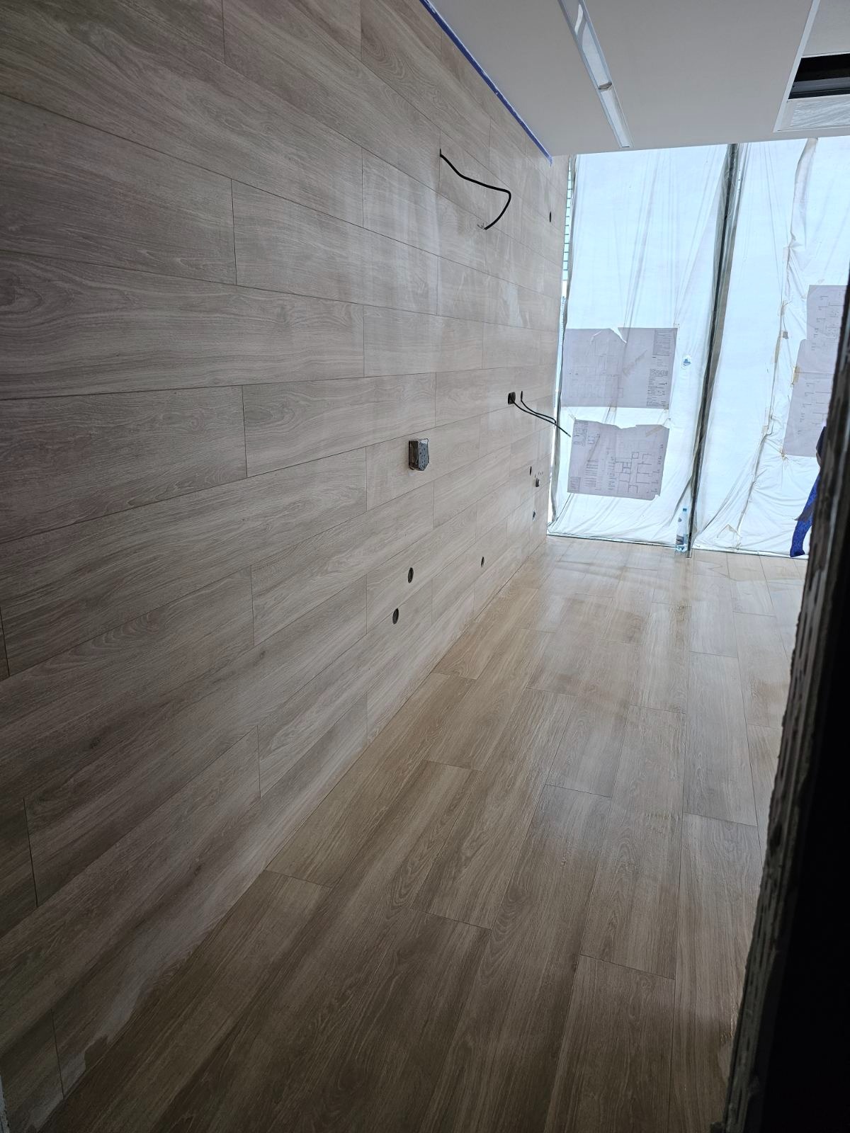 Wnętrze w trakcie prac wykończeniowych: ściana i podłoga wyłożone panelami imitującymi drewno, widoczne puszki elektryczne i otwory na kable, okno zasłonięte folią ochronną.