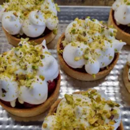 Mini tartaletki z kremem bezowym, nadzieniem owocowym i posypką pistacjową, ułożone na srebrnej tacy.