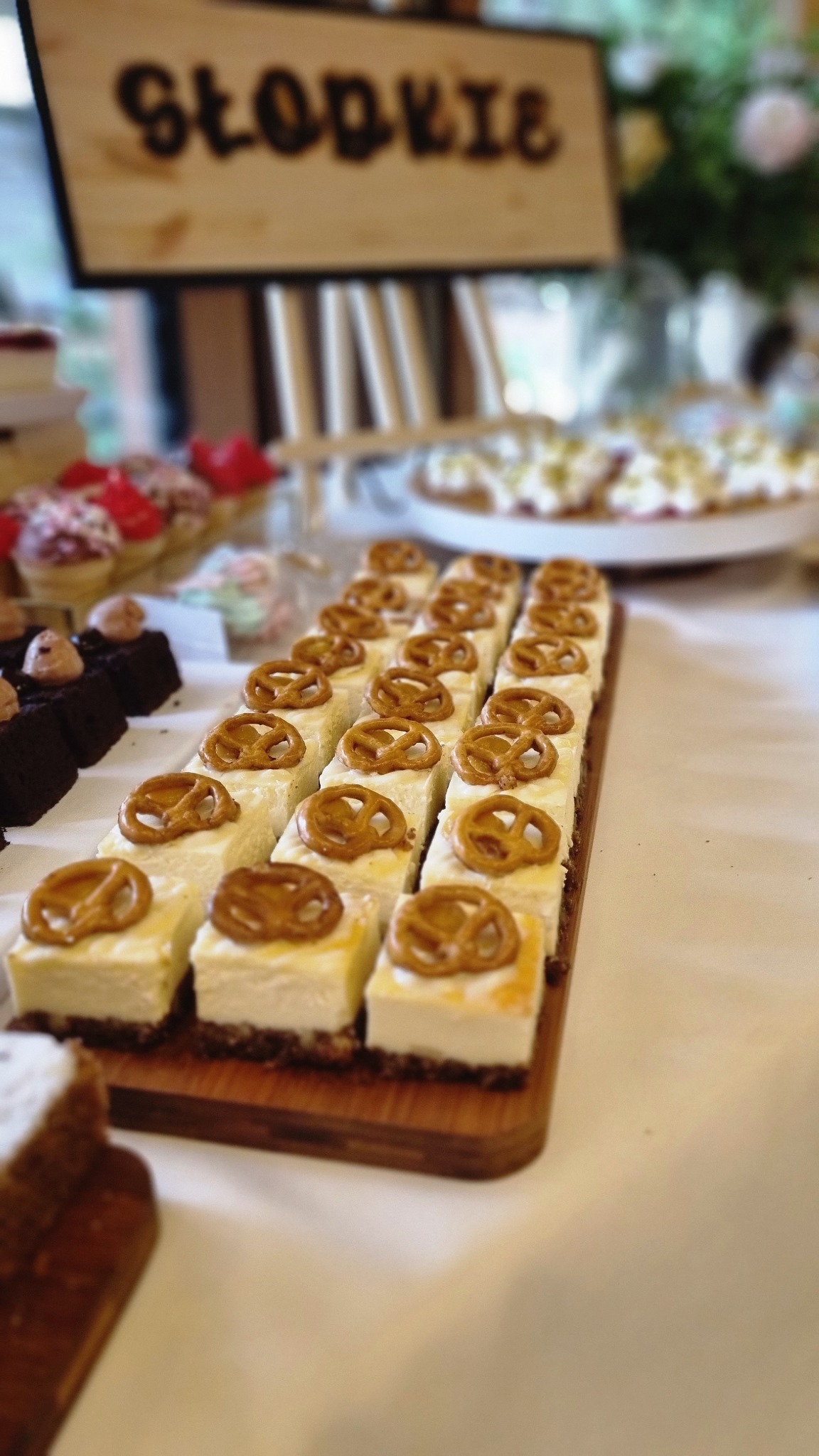 Rząd kremowych kostek sernika na ciemnym spodzie, każda udekorowana preclem, ułożone na drewnianej desce, z innymi deserami i napisem 'Słodkie' w tle.