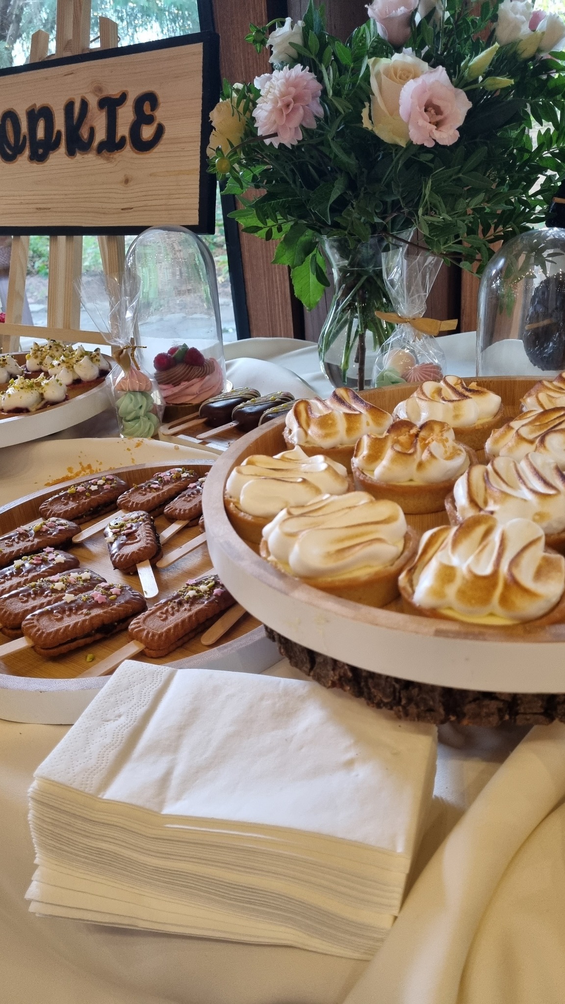 Stół z deserami: tartaletki z przypieczoną bezą, ciasteczka na patyku z czekoladą i posypką, bezy w przezroczystych opakowaniach, bukiet różowych i białych kwiatów w wazonie oraz drewniany napis...