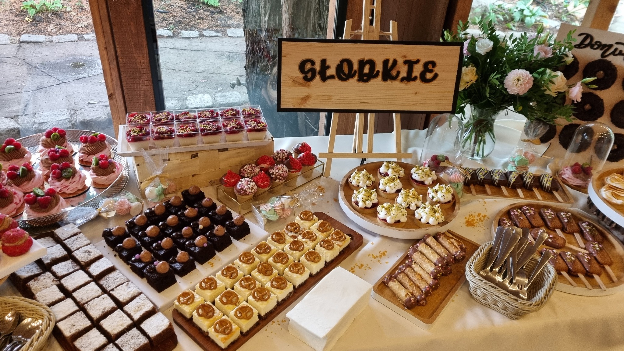 Stół z różnorodnymi deserami: babeczki z owocami, mini serniki z preclami, ciasta czekoladowe, mini tartaletki z kremem, desery w kubeczkach, w tle drewniany napis 'Słodkie' i dekoracje kwiatowe.
