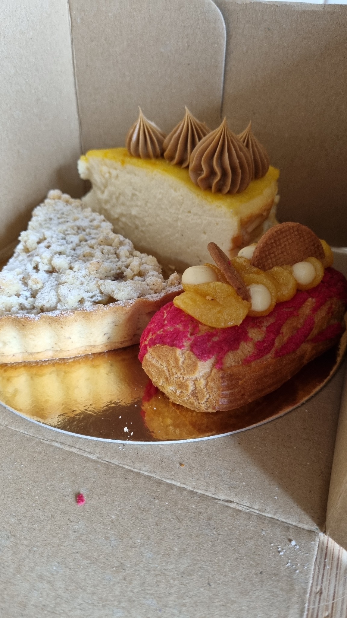 Trzy różne ciasta w kartonowym pudełku: kawałek szarlotki posypanej cukrem pudrem, kawałek sernika z karmelem i eklerka z różowym lukrem i owocami na wierzchu, ułożone na złotej tacce.