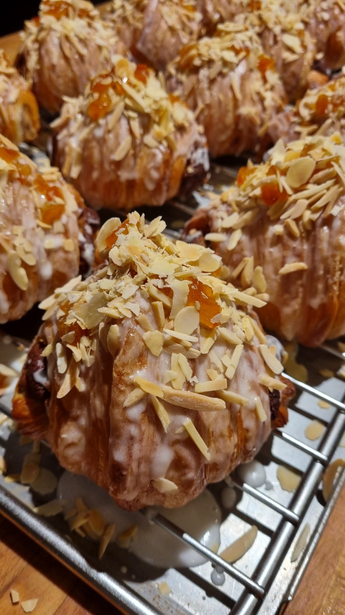 Świeżo upieczone rogaliki z nadzieniem czekoladowym, polane lukrem i posypane płatkami migdałów oraz kawałkami pomarańczowej marmolady, ułożone na metalowej kratce.