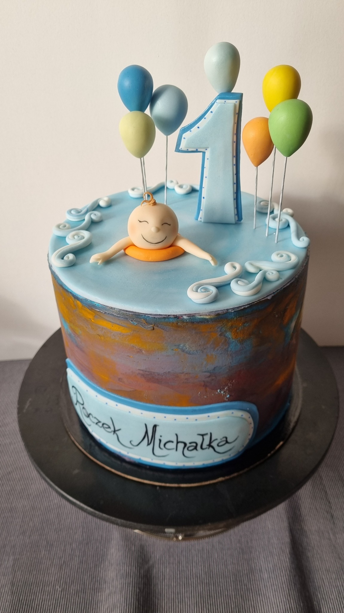 Tort urodzinowy dla roczniaka z figurką dziecka w kole ratunkowym, cyfrą '1' i balonami na błękitnym 'jeziorku' z białymi falami, spersonalizowany napisem 'Roczek Michałka' na boku z efektem ombre.