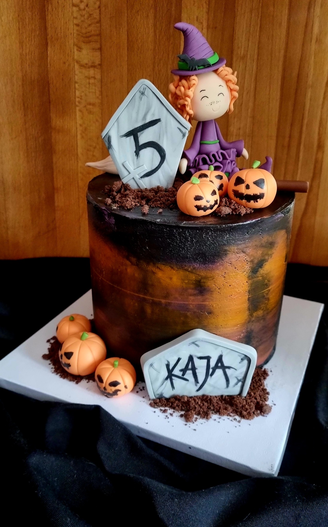 Tort urodzinowy w stylu Halloween: ciemny tort z pomarańczowymi akcentami, dekorowany figurką czarownicy, małymi dyniami i nagrobkami z napisem '5' i 'Kaja'.