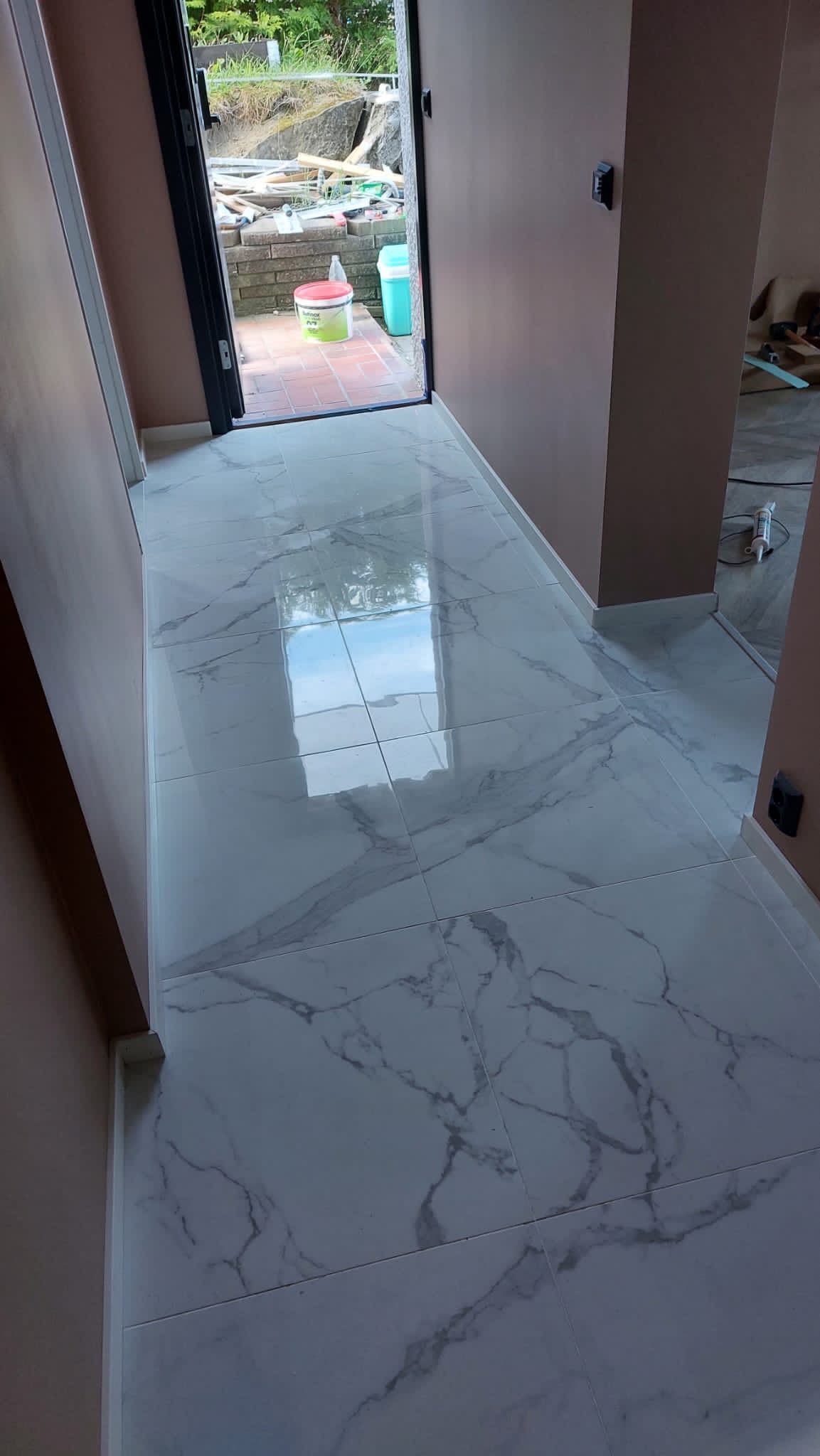 Położona, błyszcząca podłoga z płytek imitujących marmur w korytarzu, widok na otwarte drzwi i fragment ogrodu z elementami budowlanymi.