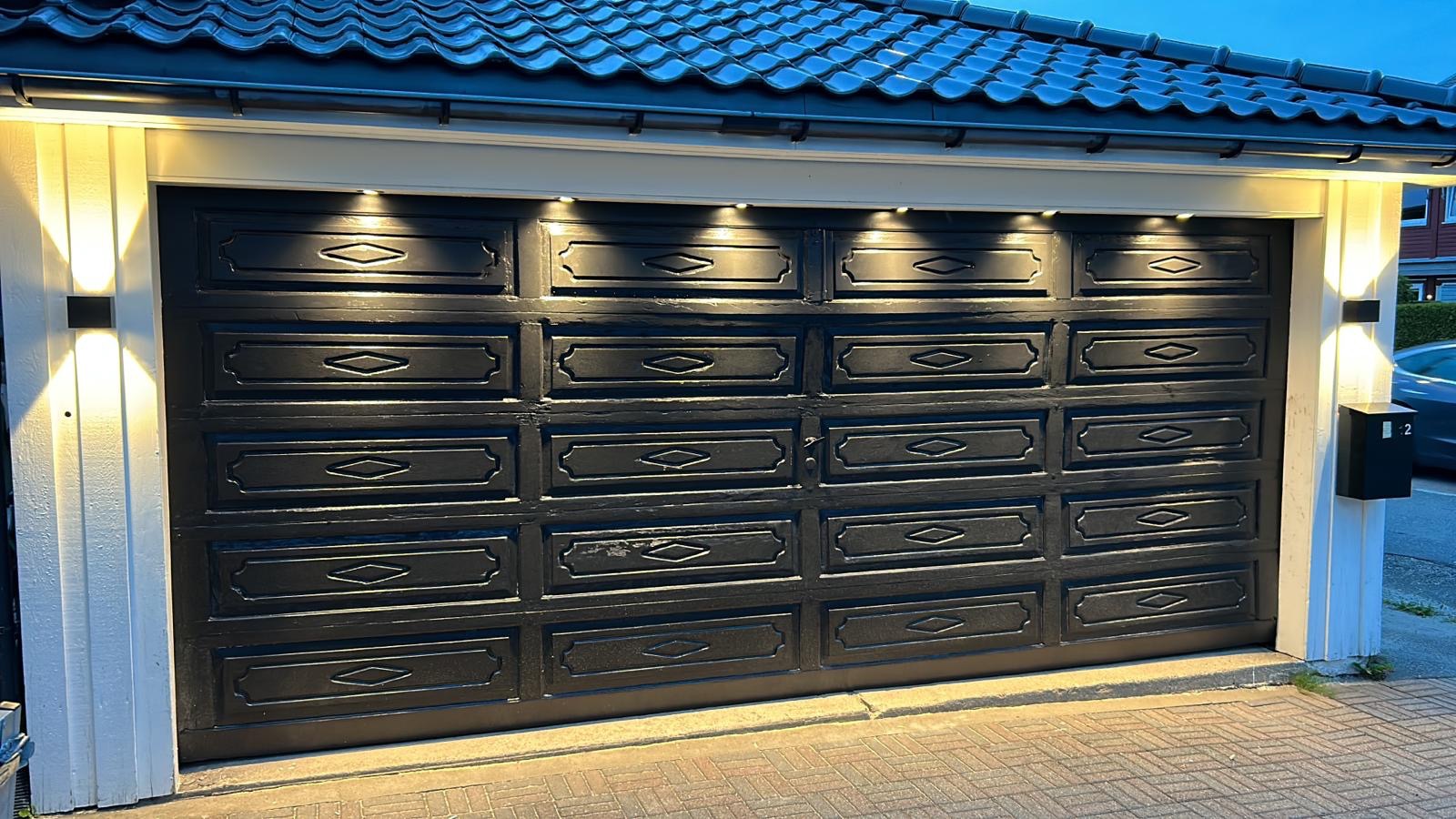 Czarna, drewniana brama garażowa z dekoracyjnymi panelami, oświetlona od góry wbudowanymi lampkami i dwoma zewnętrznymi kinkietami, widoczna skrzynka pocztowa po prawej stronie.