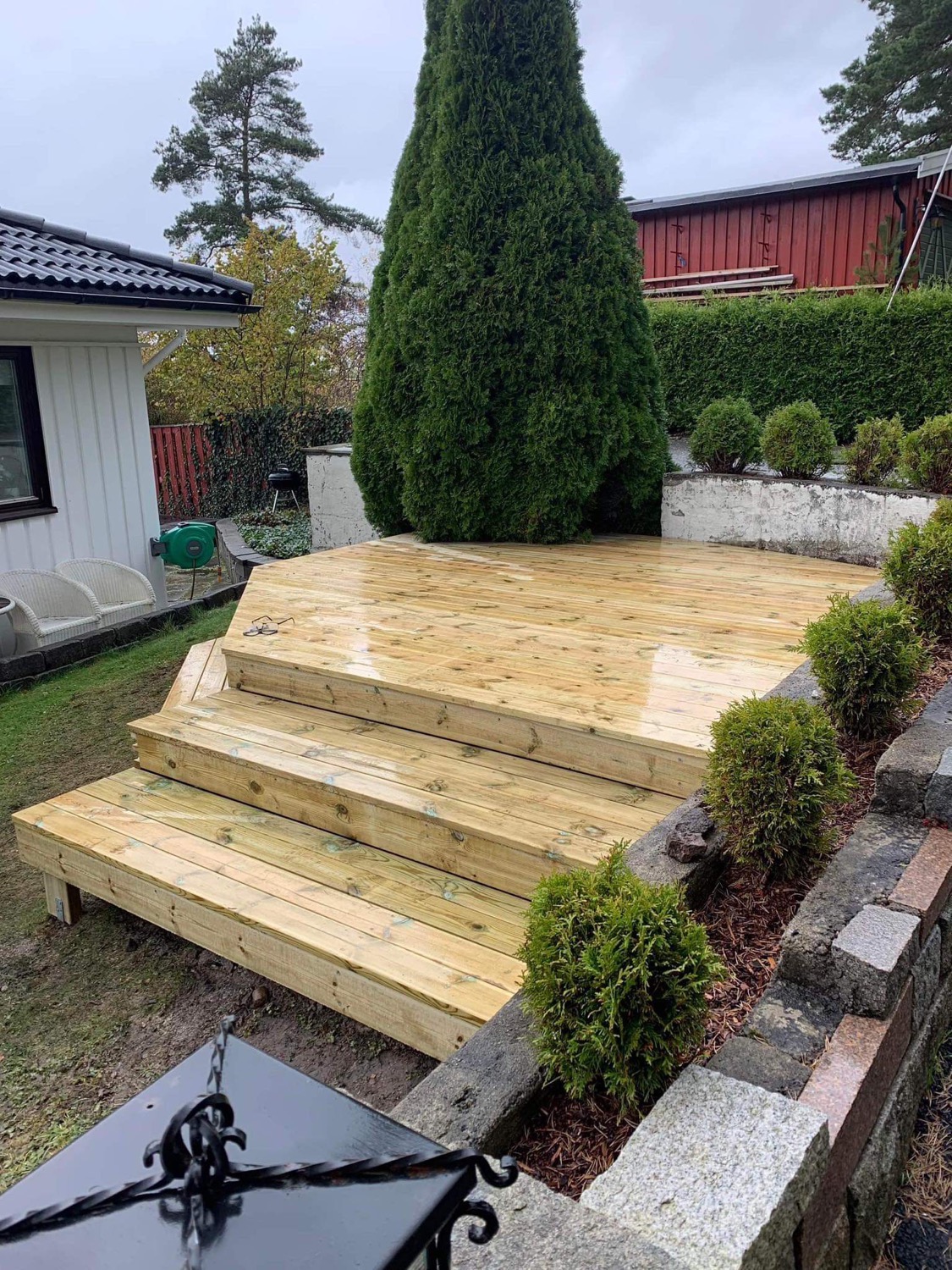 Nowo wybudowany, wielopoziomowy taras z jasnego drewna z trzema stopniami, otoczony niską ścianką z kamienia i krzewami, z widocznym fragmentem domu w tle.