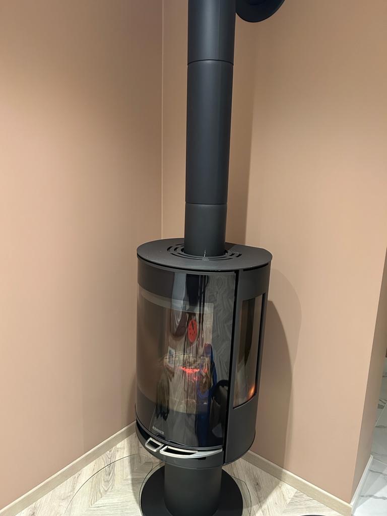 Czarny, nowoczesny piec wolnostojący z widocznym płomieniem, ustawiony w rogu pokoju z beżowymi ścianami i jasną podłogą imitującą drewno.