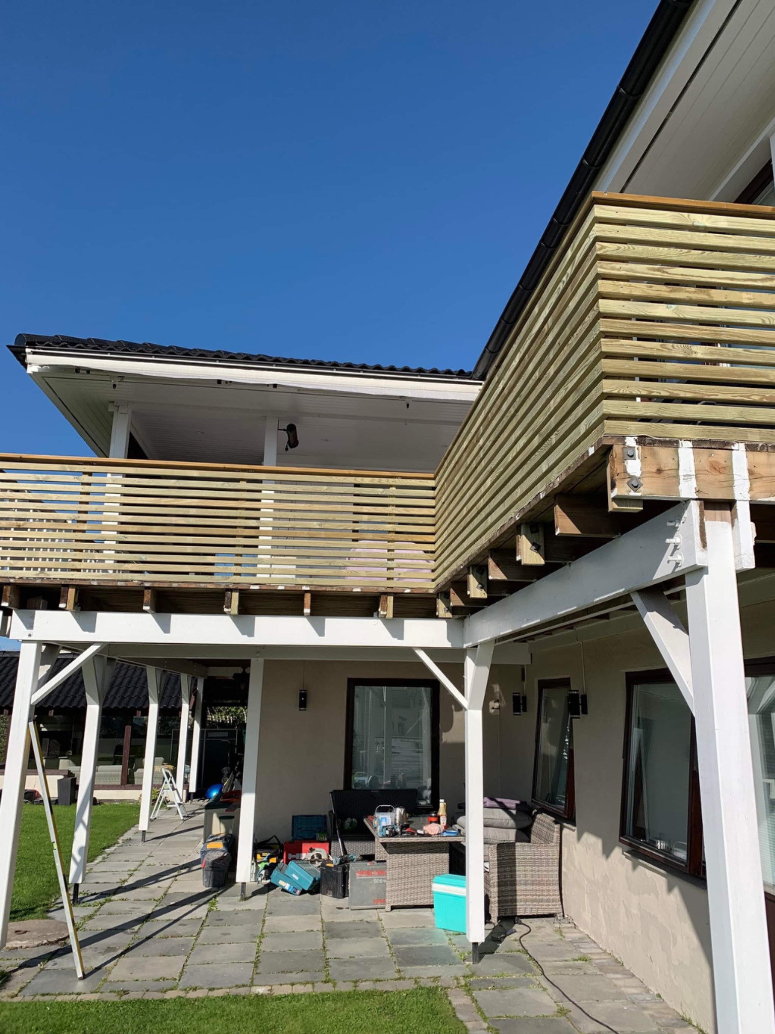 Nowo wybudowany drewniany balkon z poziomymi deskami, widok z dołu na tle błękitnego nieba i części domu. Na tarasie widoczne narzędzia i meble ogrodowe.