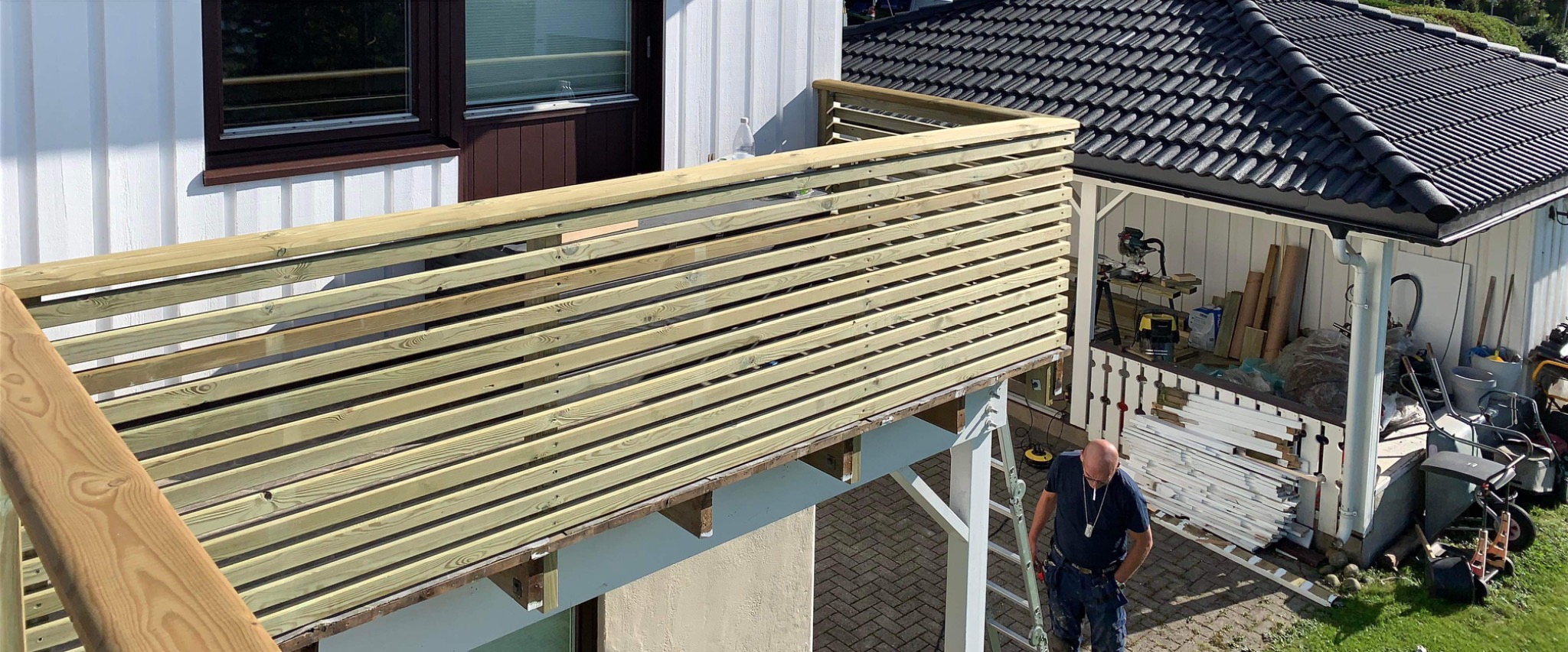 Montaż drewnianej balustrady na balkonie domu jednorodzinnego, widok z góry na pracownika na drabinie, z widocznymi elementami konstrukcyjnymi i narzędziami w tle.