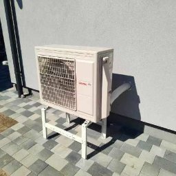 DanLuk Daniel Łukaszewski - Zewnętrzna jednostka klimatyzacji Atlantic na białym stelażu, zamontowana przy jasnej ścianie budynku, widoczna rura odprowadzająca skropliny.