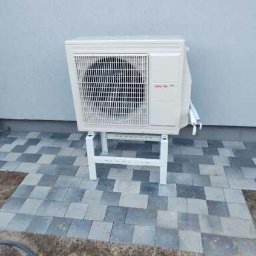 DanLuk Daniel Łukaszewski - Biała jednostka zewnętrzna pompy ciepła marki Atlantic, umieszczona na białym stojaku na szaro-niebieskim brukowanym podłożu, z szarą ścianą w tle.