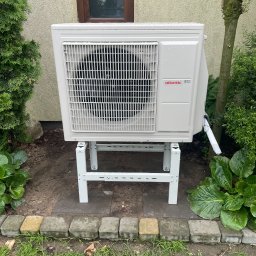 DanLuk Daniel Łukaszewski - Biała jednostka zewnętrzna pompy ciepła Atlantic R32 na metalowym stelażu, umieszczona w ogrodzie przy ścianie budynku, z widocznymi roślinami i kostką brukową.