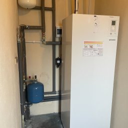 DanLuk Daniel Łukaszewski - Biała pompa ciepła Atlantic w pomieszczeniu technicznym, z widocznymi rurami, zbiornikami wyrównawczymi i manometrami.