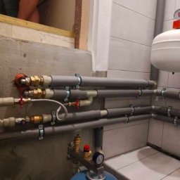 DanLuk Daniel Łukaszewski - Instalacja grzewcza z rurami w izolacji, zaworami i manometrami, zamontowana przy ścianie z płytkami. Widoczny zbiornik wyrównawczy.