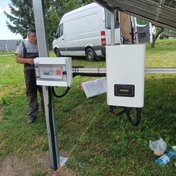 DanLuk Daniel Łukaszewski - Instalator w kombinezonie roboczym przy skrzynce z bezpiecznikami i inwerterem, montowanych na metalowej konstrukcji na trawie, z białym vanem w tle.