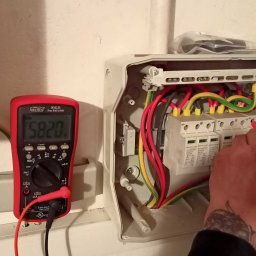 DanLuk Daniel Łukaszewski - Pomiar napięcia na zabezpieczeniach przepięciowych w rozdzielnicy elektrycznej, widoczny multimetr Brymen BM235 z sondami w rękach w czerwonych rękawicach roboczych.