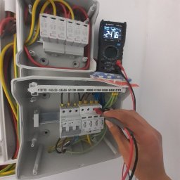 DanLuk Daniel Łukaszewski - Sprawdzanie napięcia w rozdzielnicy za pomocą multimetru cyfrowego, widoczne bezpieczniki i przewody w kolorach czerwonym, żółtym i zielonym.