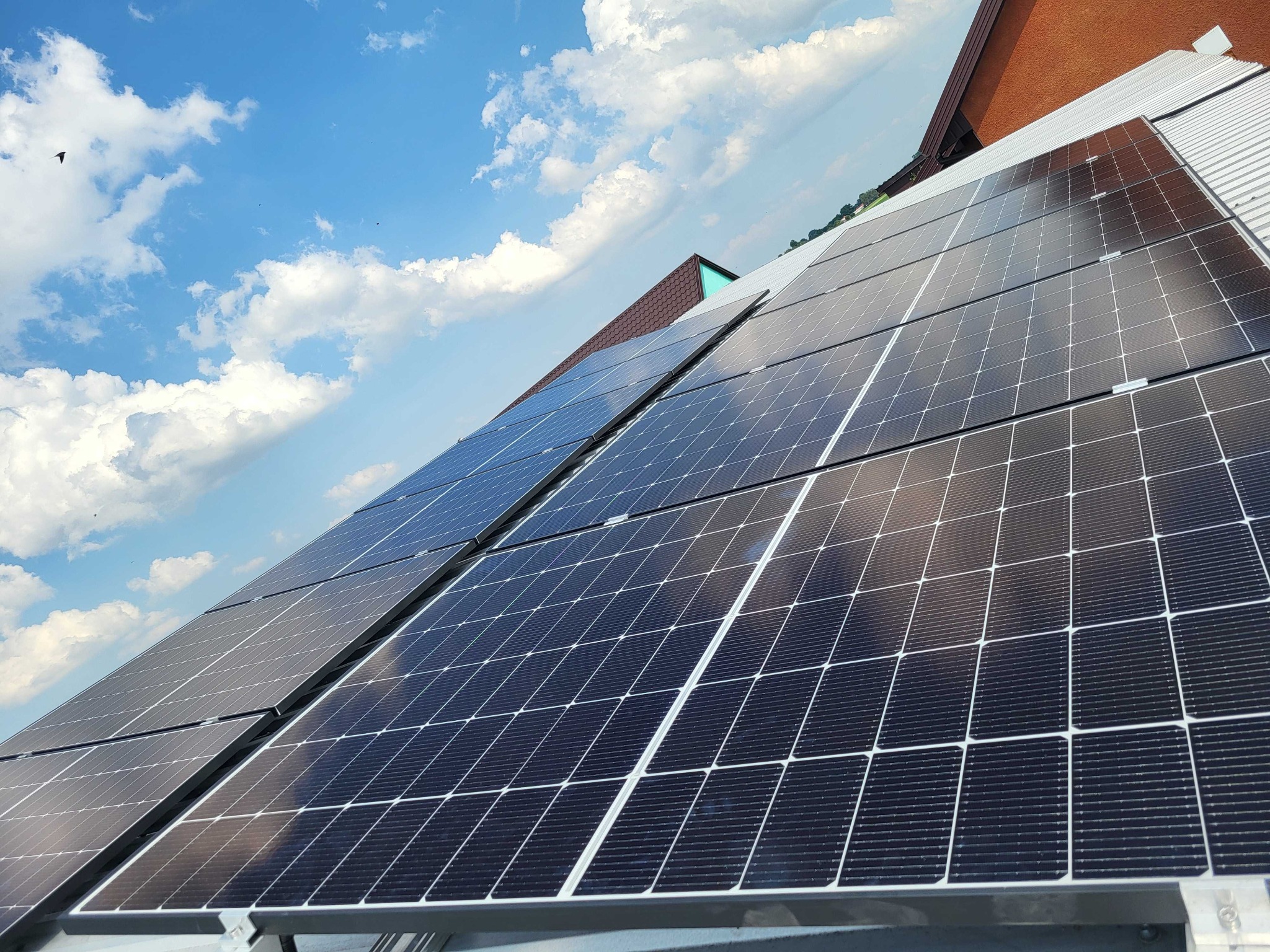 Panele słoneczne na dachu budynku z blachy trapezowej, widoczne mocowania i elementy konstrukcyjne, na tle błękitne niebo z białymi chmurami i fragment dachu sąsiedniego budynku.