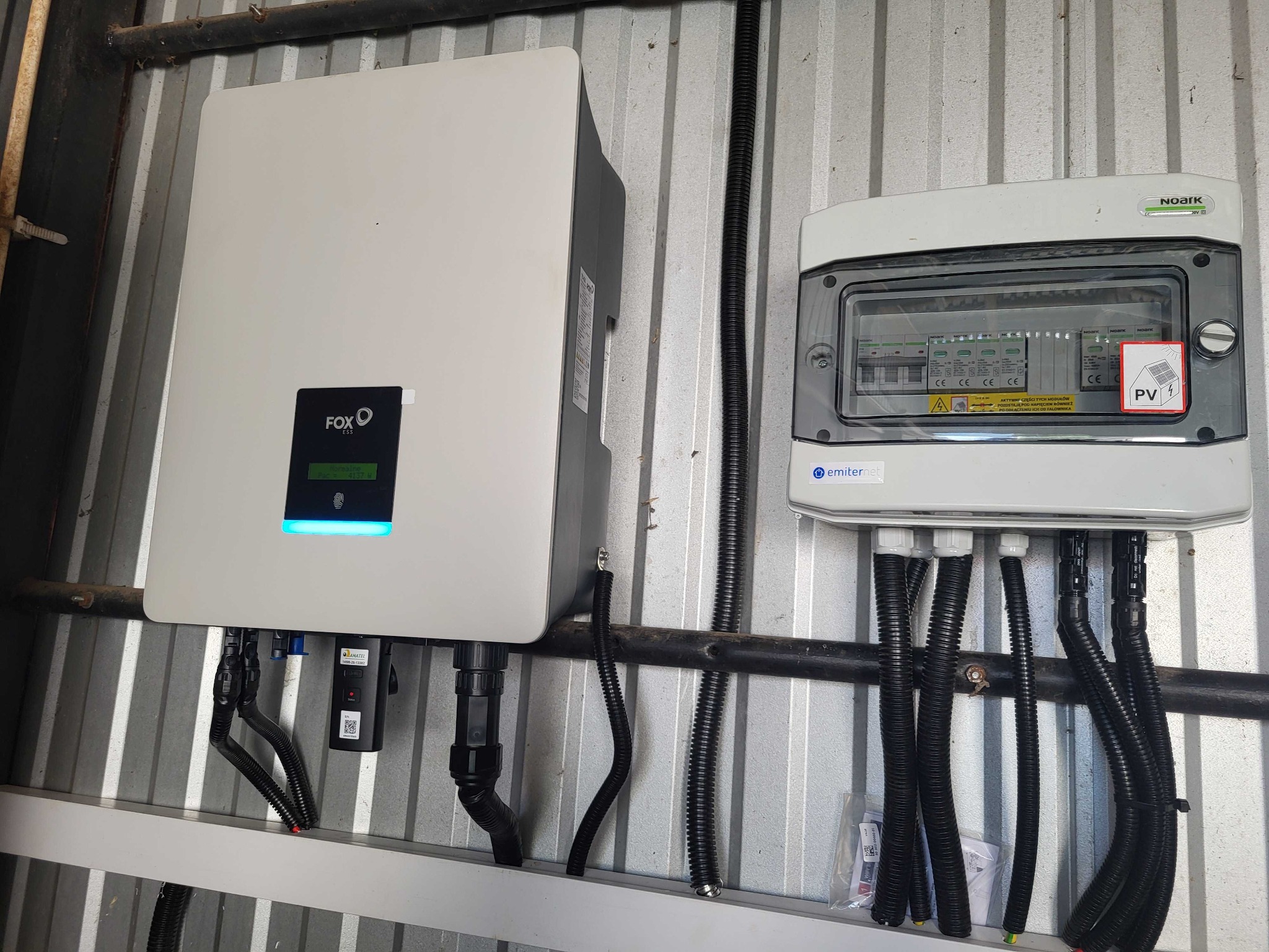 Zamontowany inwerter Fox ESS z wyświetlaczem i skrzynka zabezpieczeń NOARK z naklejką PV4 na blaszanym podłożu, widoczne czarne peszle z kablami.