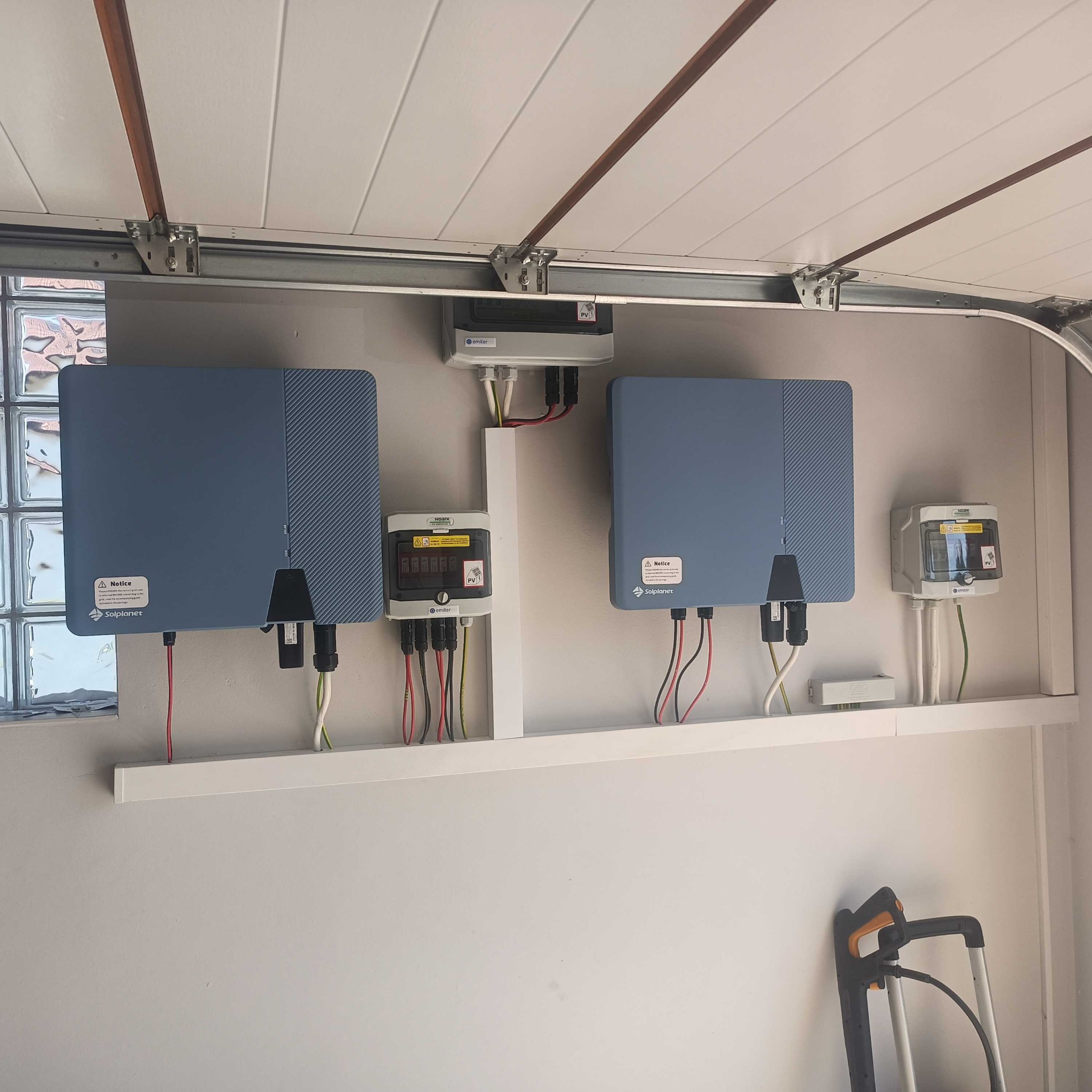 Dwa falowniki Solplanet i rozdzielnice Noark zamontowane na ścianie w garażu, widoczne okno z luksferami i częściowo złożona drabina.