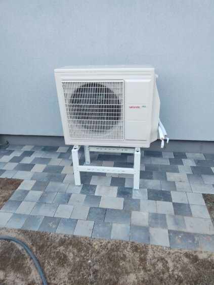 Biała jednostka zewnętrzna pompy ciepła marki Atlantic, umieszczona na białym stojaku na szaro-niebieskim brukowanym podłożu, z szarą ścianą w tle.