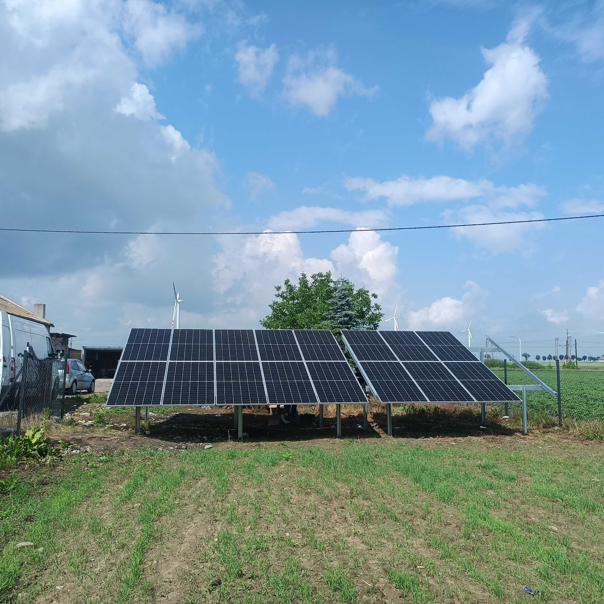 Dwa rzędy paneli słonecznych na metalowej konstrukcji, na tle błękitnego nieba z chmurami i oddalonych wiatraków, widoczne budynki gospodarcze i samochody.