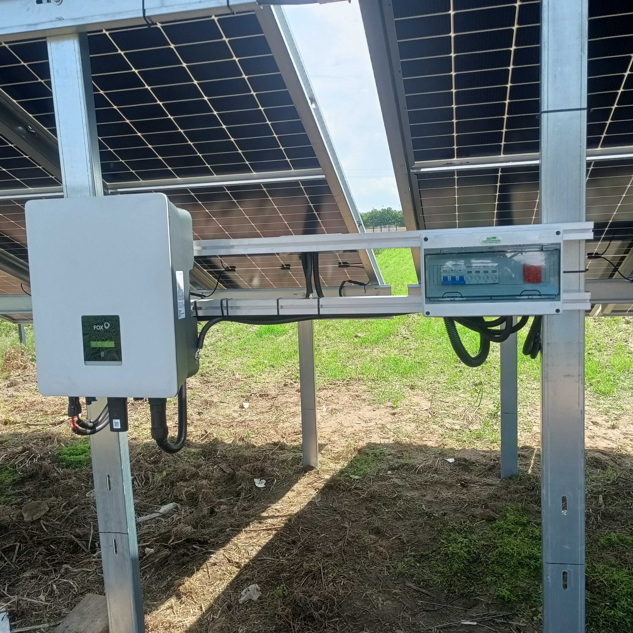 Instalacja fotowoltaiczna: widok na inwerter FoxESS, skrzynkę zabezpieczeń i panele słoneczne na metalowej konstrukcji wsporczej, z zielonym trawiastym tłem.