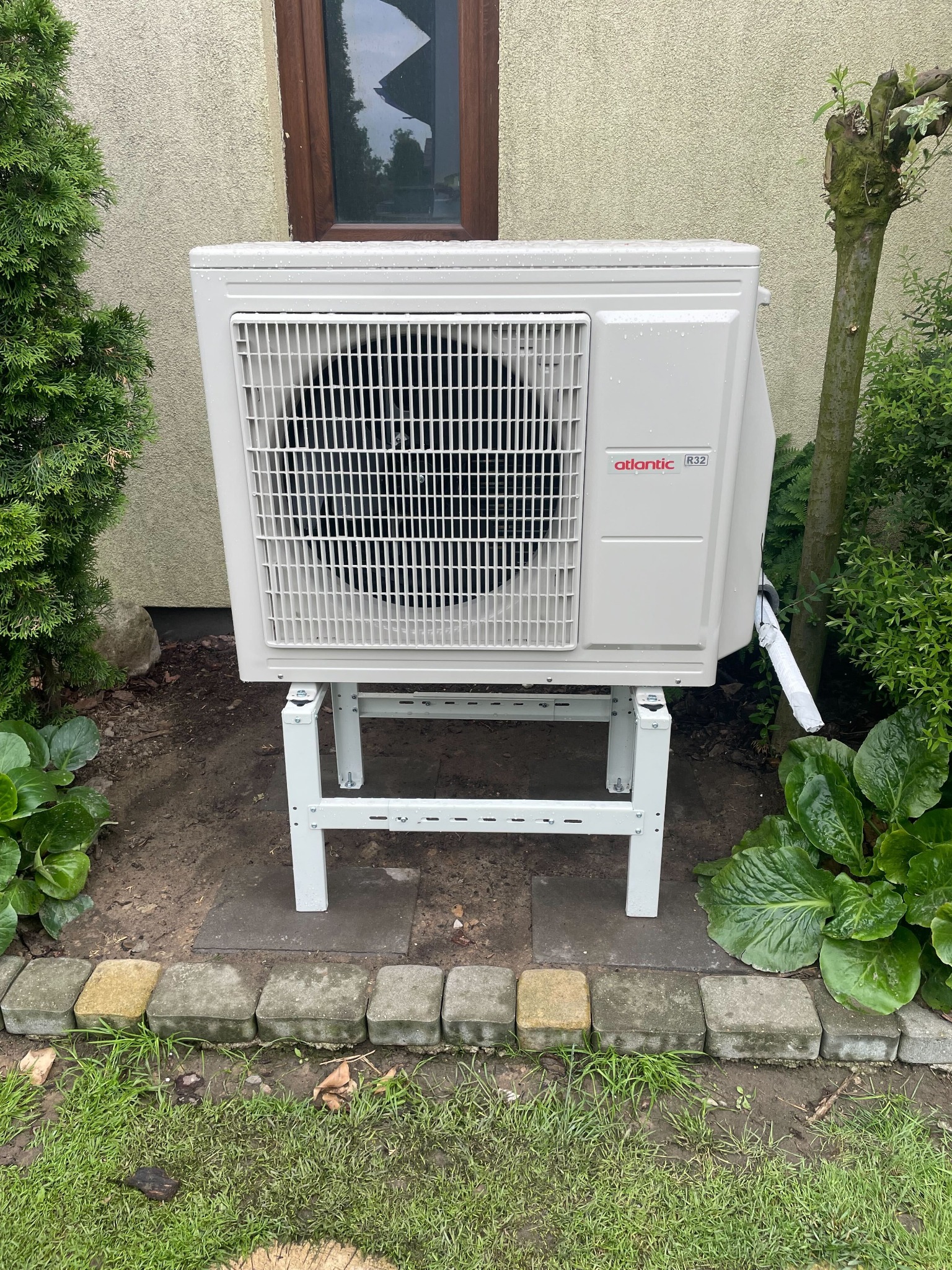 Biała jednostka zewnętrzna pompy ciepła Atlantic R32 na metalowym stelażu, umieszczona w ogrodzie przy ścianie budynku, z widocznymi roślinami i kostką brukową.