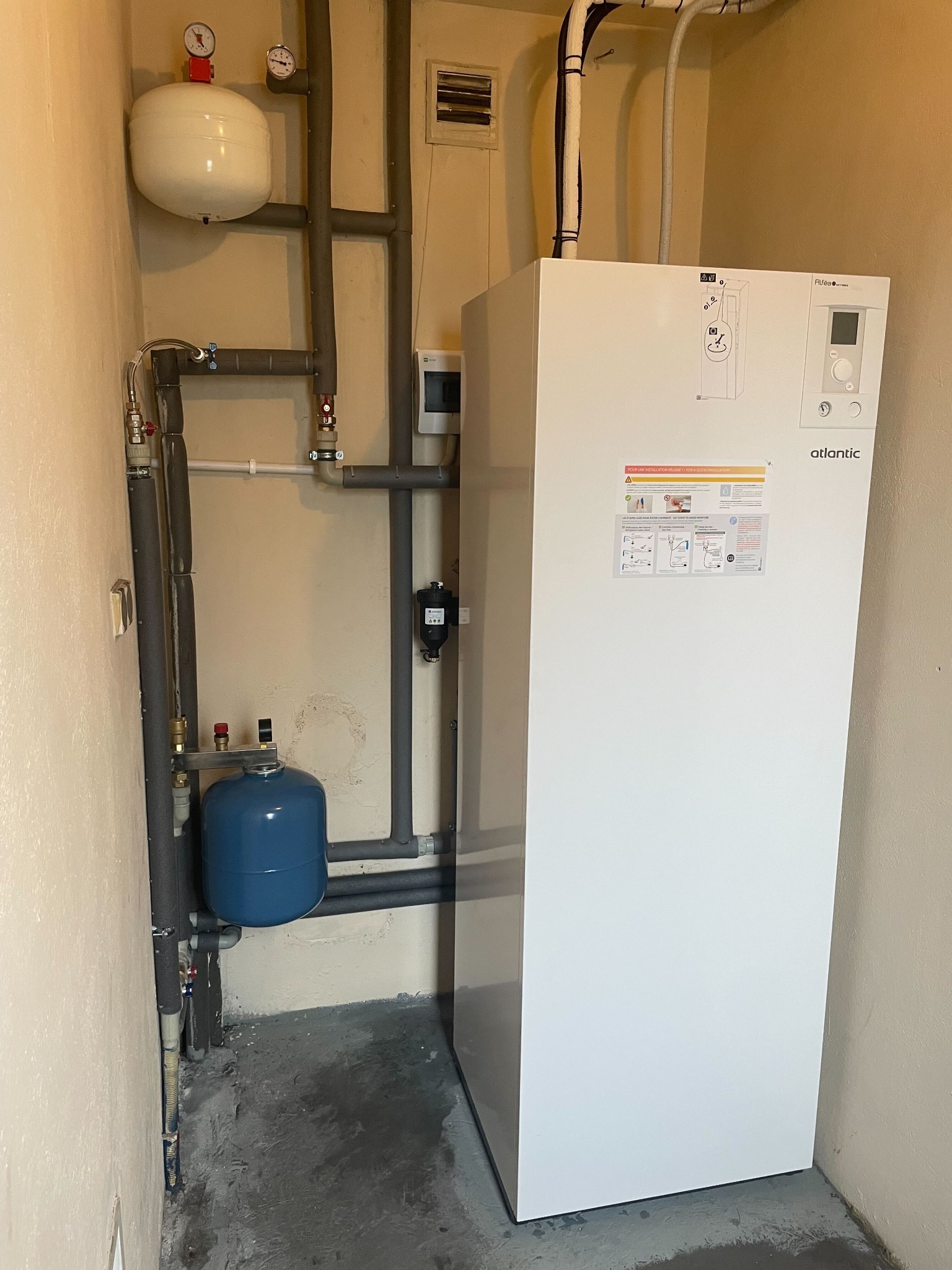 Biała pompa ciepła Atlantic w pomieszczeniu technicznym, z widocznymi rurami, zbiornikami wyrównawczymi i manometrami.