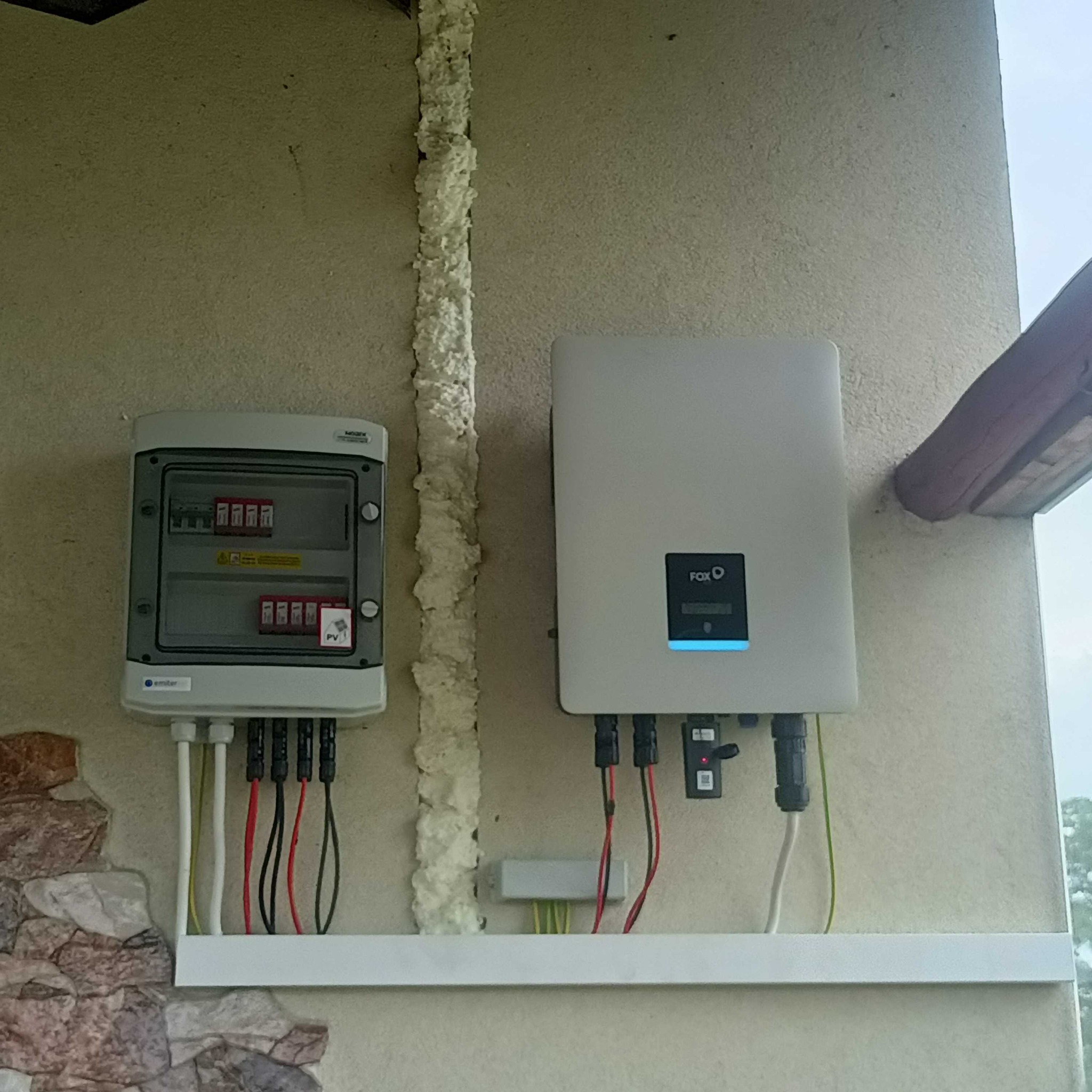 Zamontowana na zewnątrz skrzynka rozdzielcza i inwerter FoxESS z niebieskim podświetleniem, widoczne kable i pianka montażowa uszczelniająca przestrzeń.