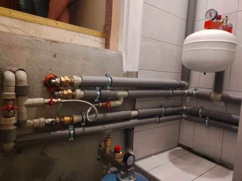 Instalacja grzewcza z rurami w izolacji, zaworami i manometrami, zamontowana przy ścianie z płytkami. Widoczny zbiornik wyrównawczy.