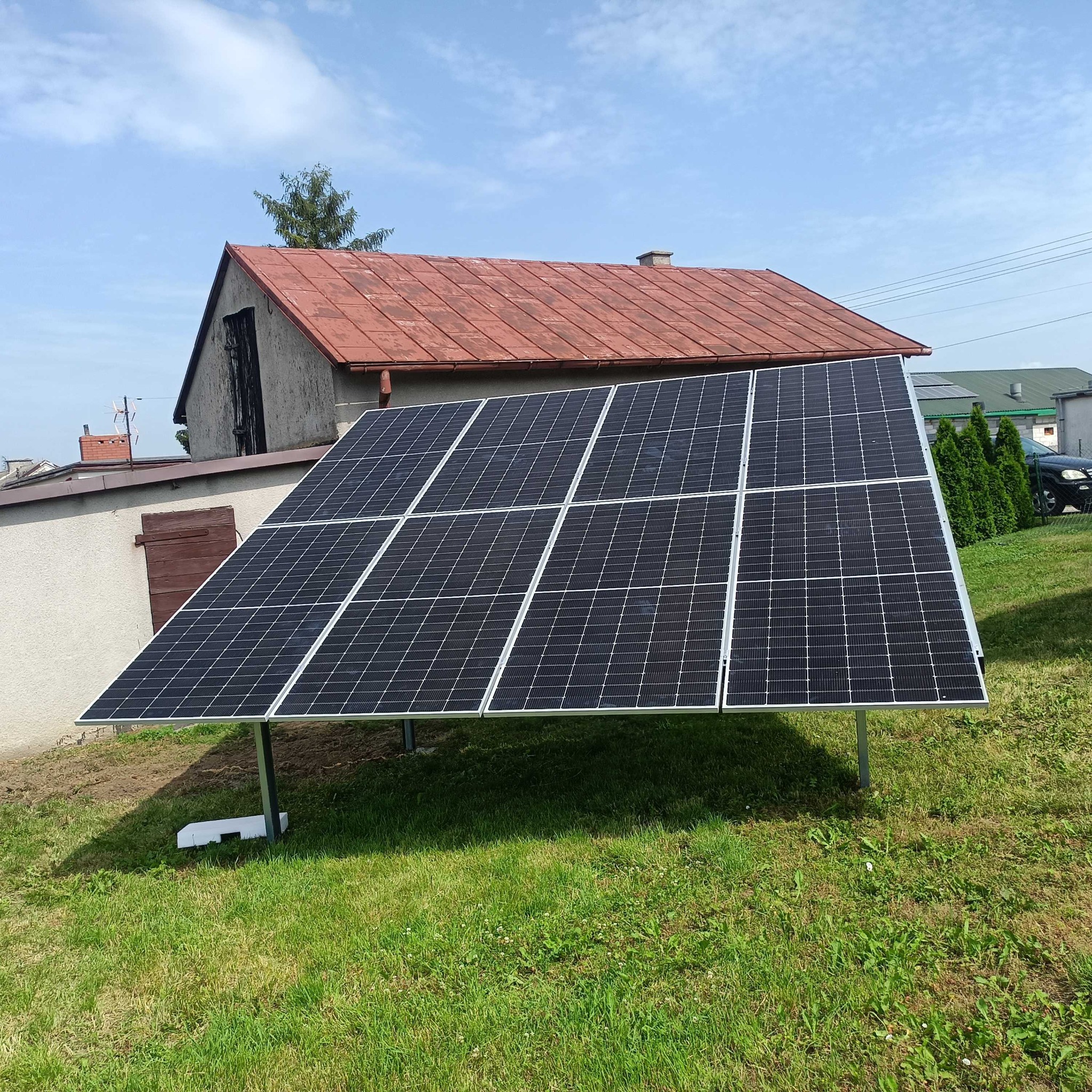 Panel słoneczny na trawie przed budynkiem z czerwoną dachówką, widoczne podparcie i osprzęt.