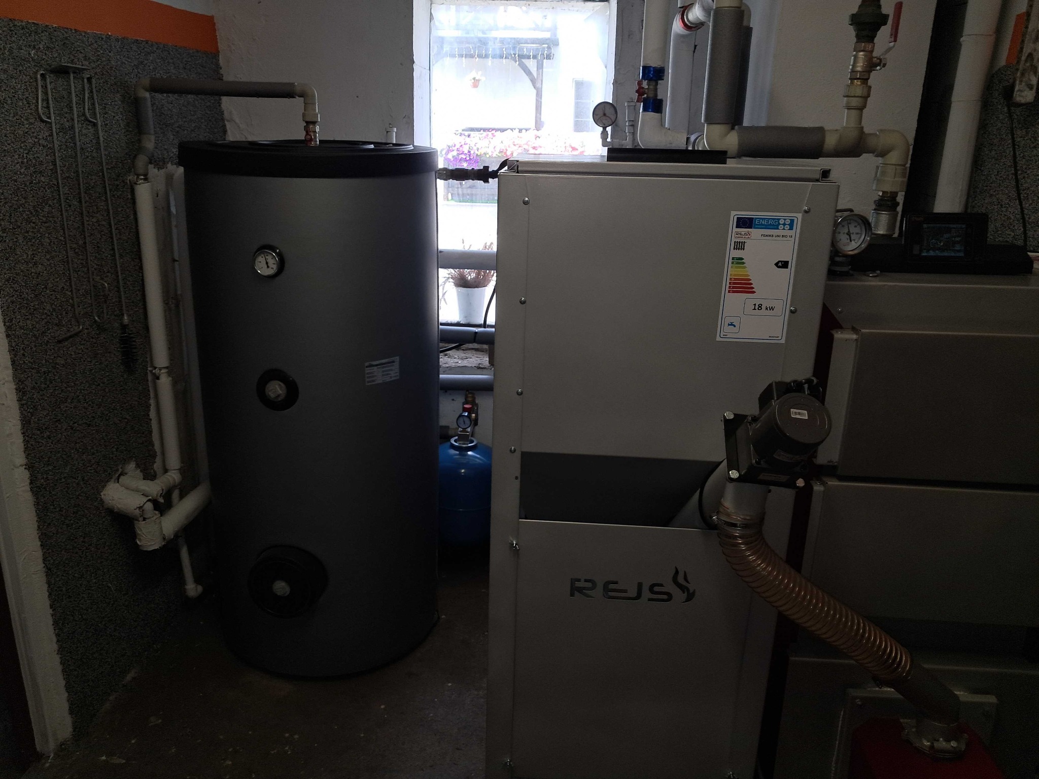 Szary zbiornik akumulacyjny ciepła obok kotła grzewczego z widocznym logo REJS, instalacja w kotłowni z oknem.